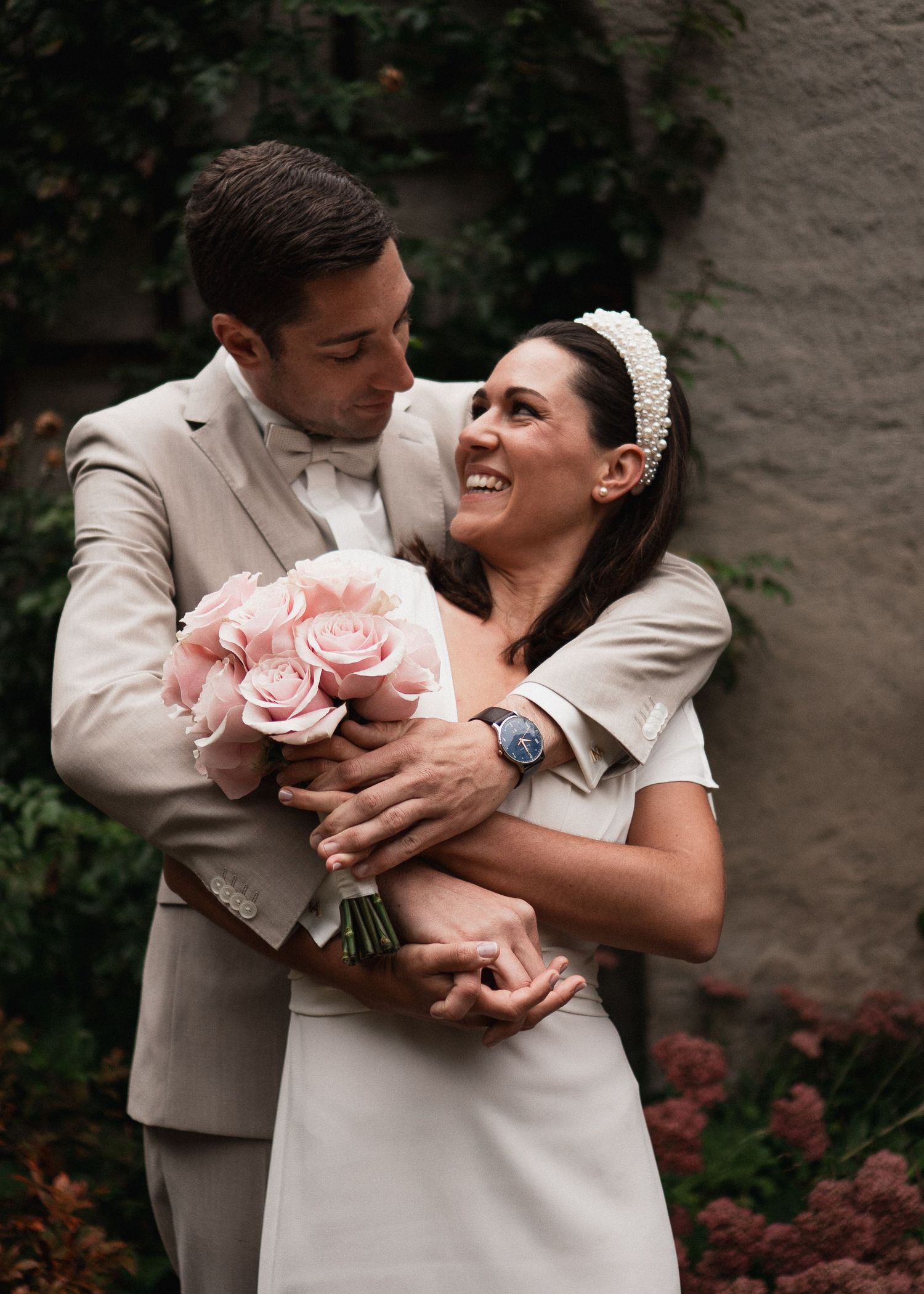 Romantisches Brautpaarfotografie im Sichtungsgarten Weihenstephan: Bräutigam umarmt seine lachende Braut mit rosa Rosenstrauß nach der stand
