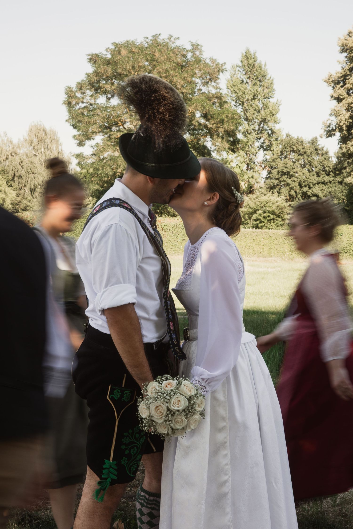Trachten-Hochzeit in Bad Birnbach: Brautpaar küsst sich innig, während Hochzeitsgäste in Tracht verschwommen an ihnen vorbeilaufen.
