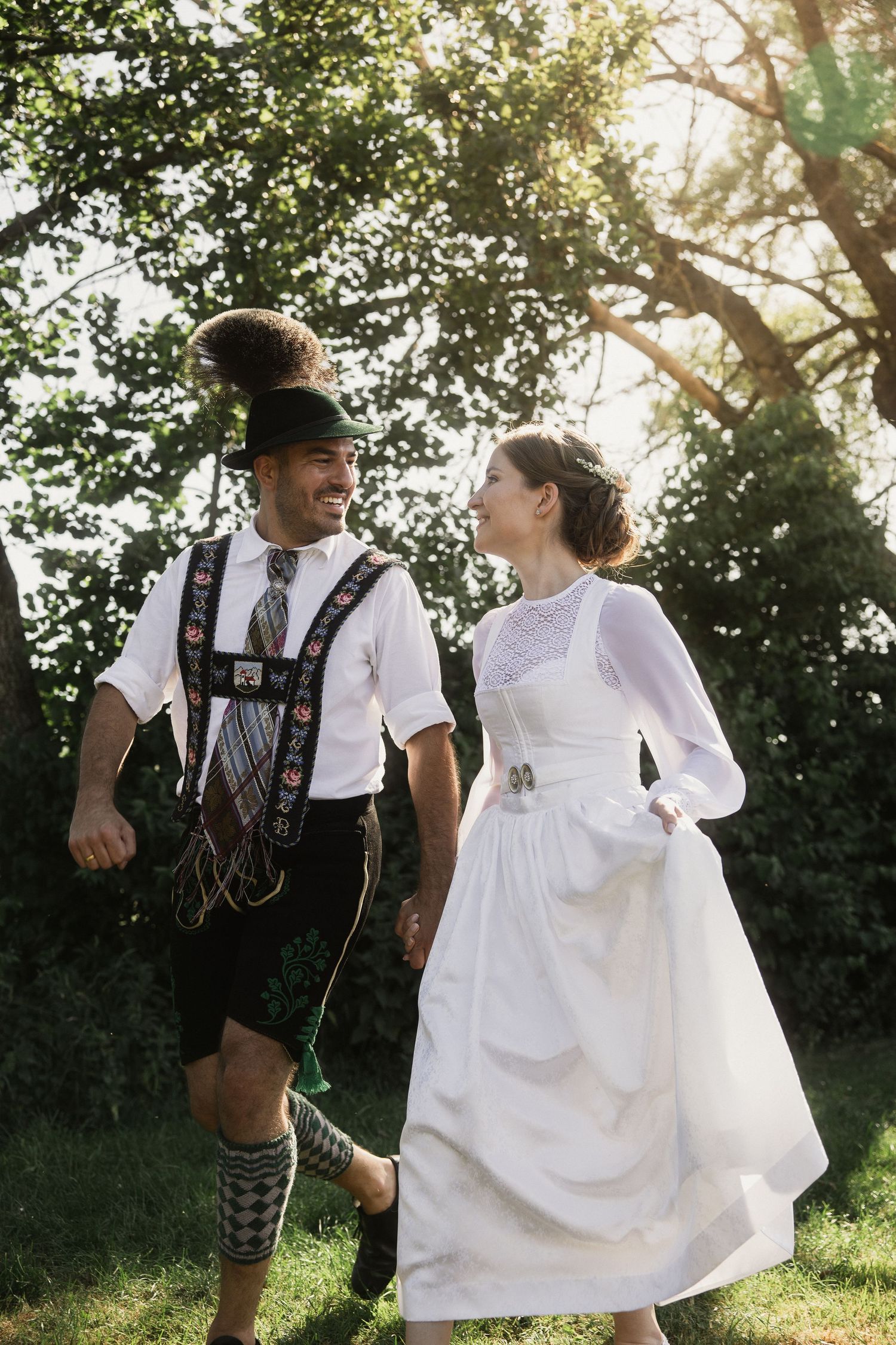 Trachten-Hochzeit in Bad Birnbach: Brautpaar in Dirndl und Lederhosen läuft lachend Hand in Hand durchs Grüne im Gegenlicht.