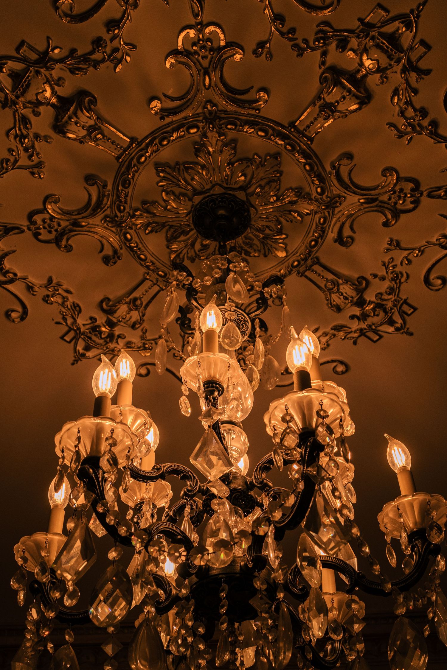 Ornate vintage crystal chandelier with warm glowing lights illuminates an elegant ceiling with baroque-style decorative details.