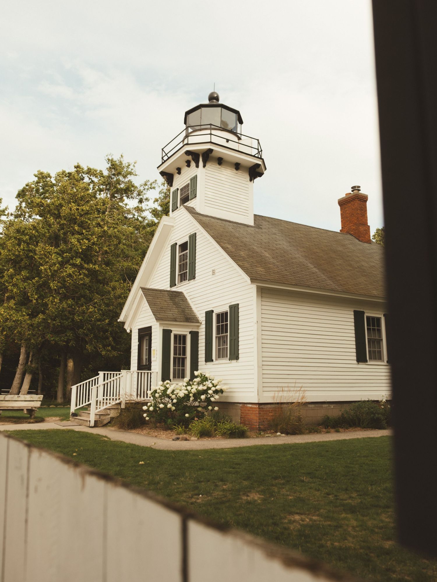 White lighthouse keeper's house with black shutters, tower, and red brick chimney surrounded by green trees and lawn.