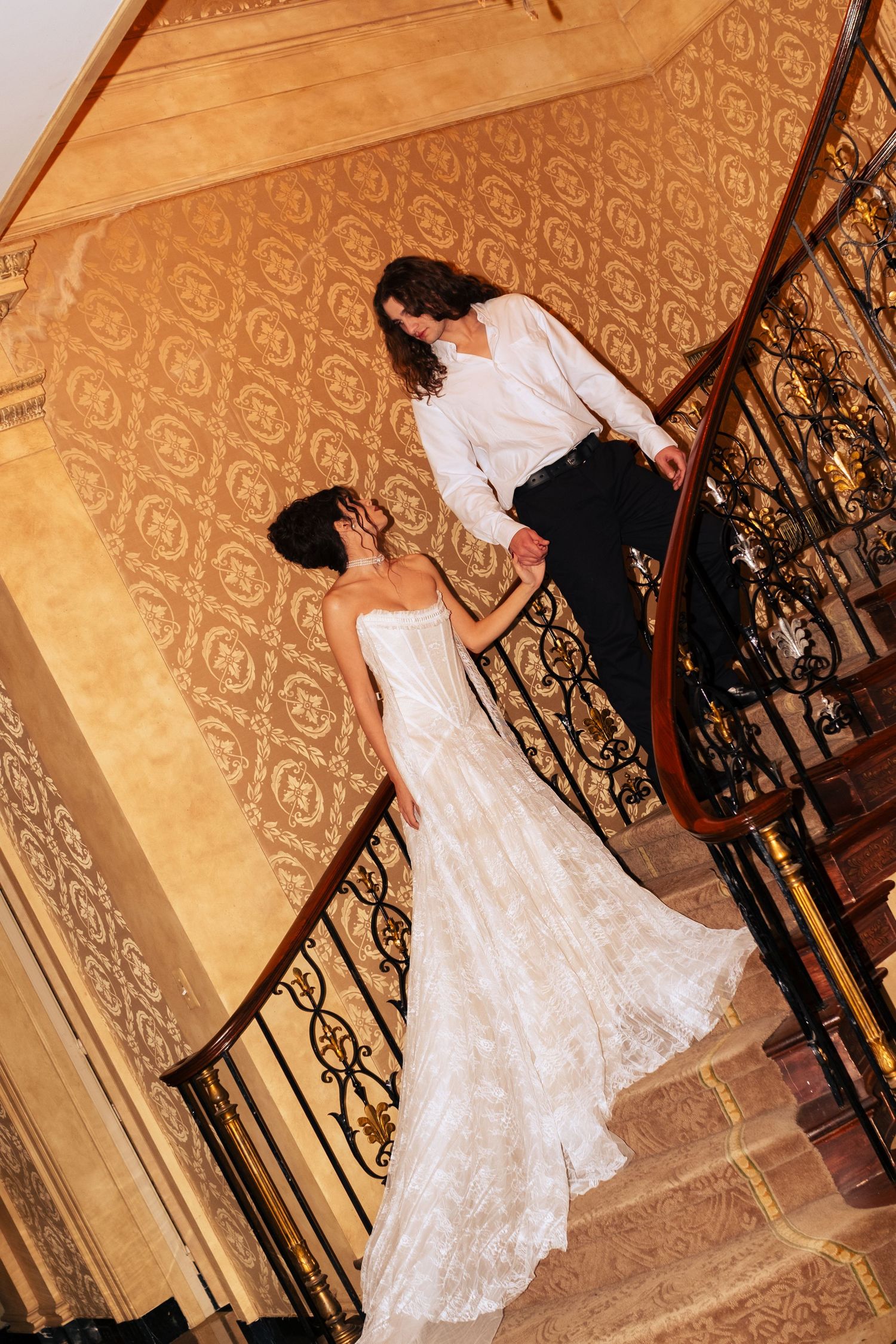 A bride in a flowing white lace gown descends an elegant staircase with ornate wrought iron railings and patterned walls.