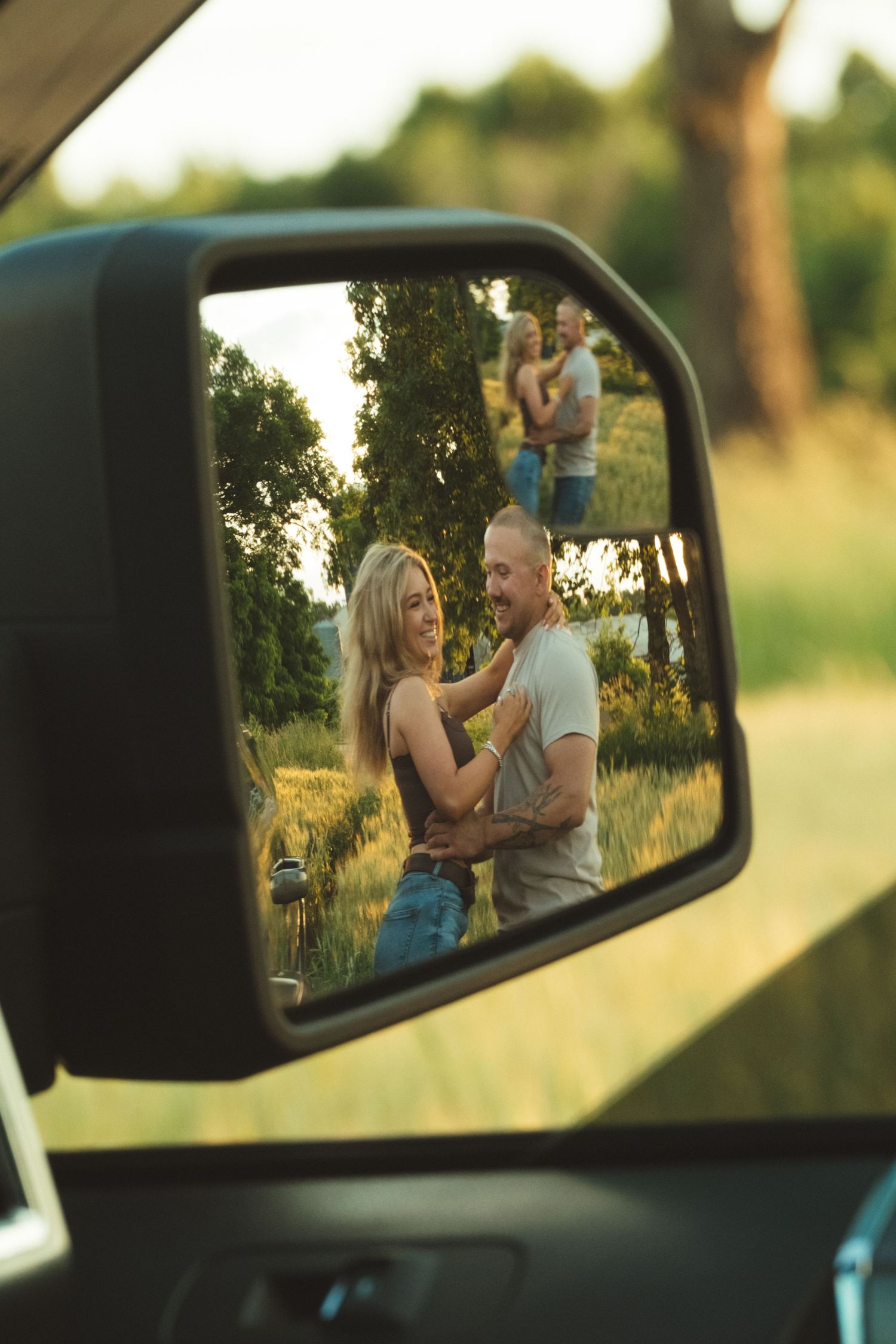 Couple embracing in a field reflected in a car side mirror on a sunny day.