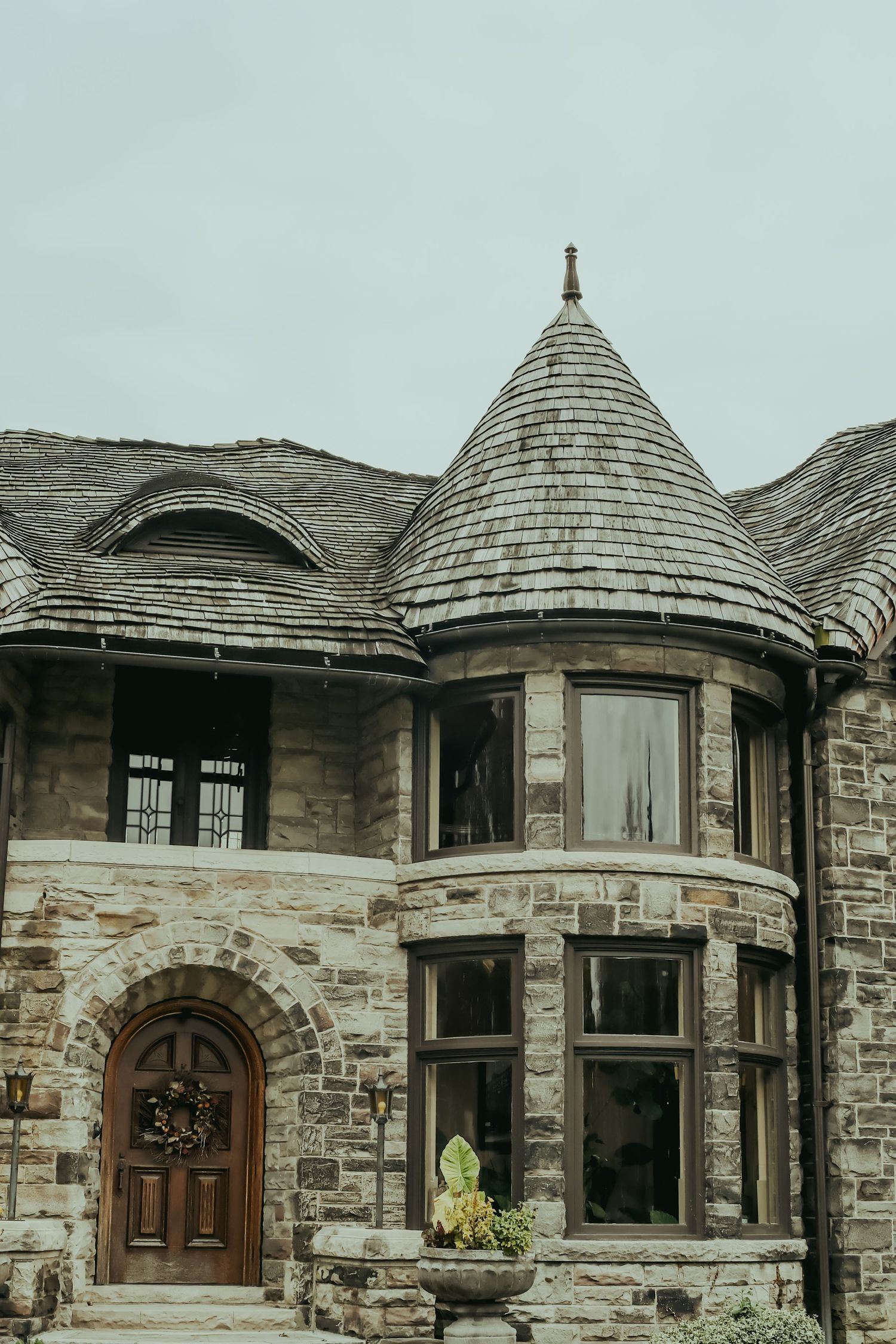 Historic stone mansion with conical turret, arched wooden door, and slate roof under overcast sky.