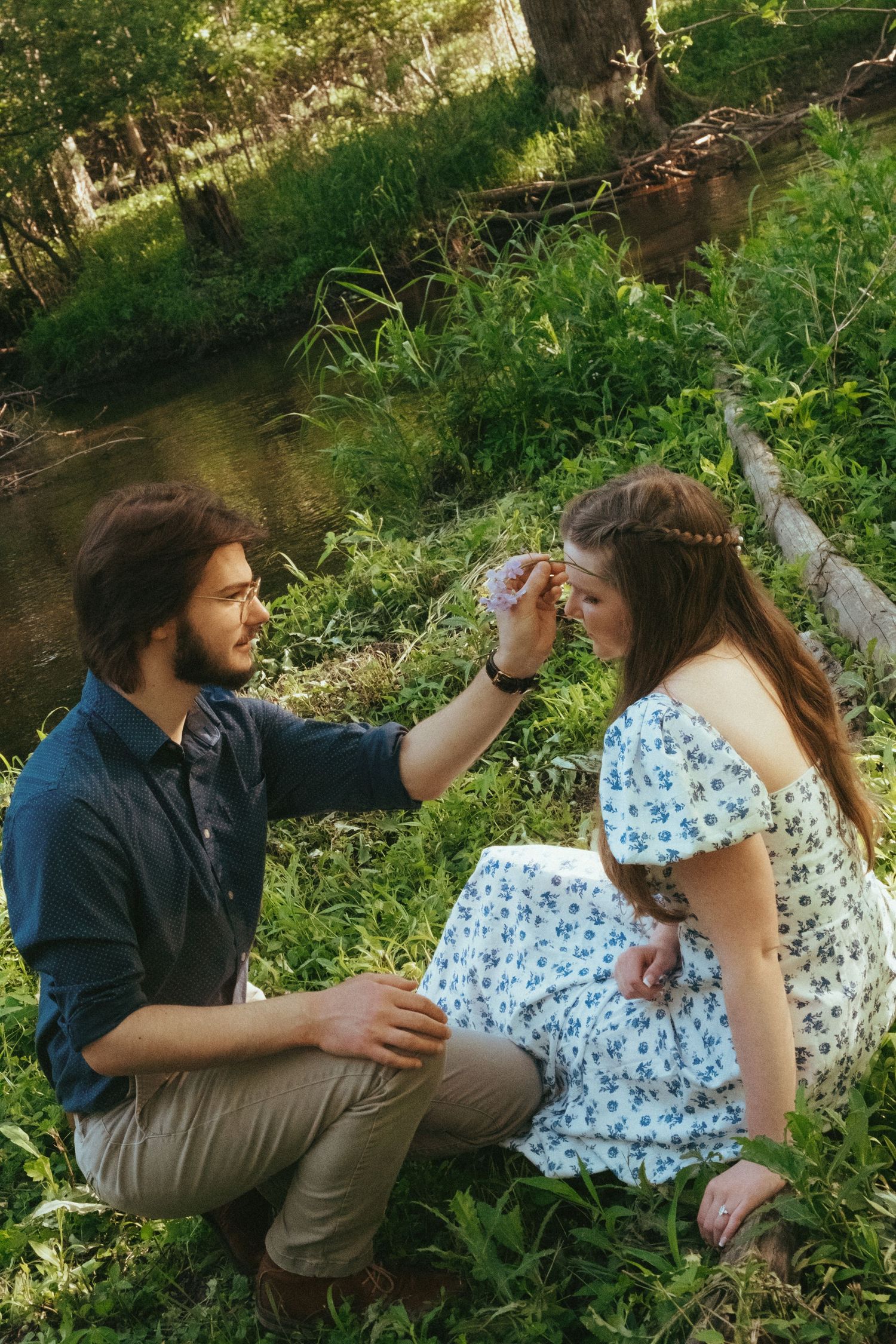 A couple sits together in a lush green forest, man tenderly touching womans face near a stream.