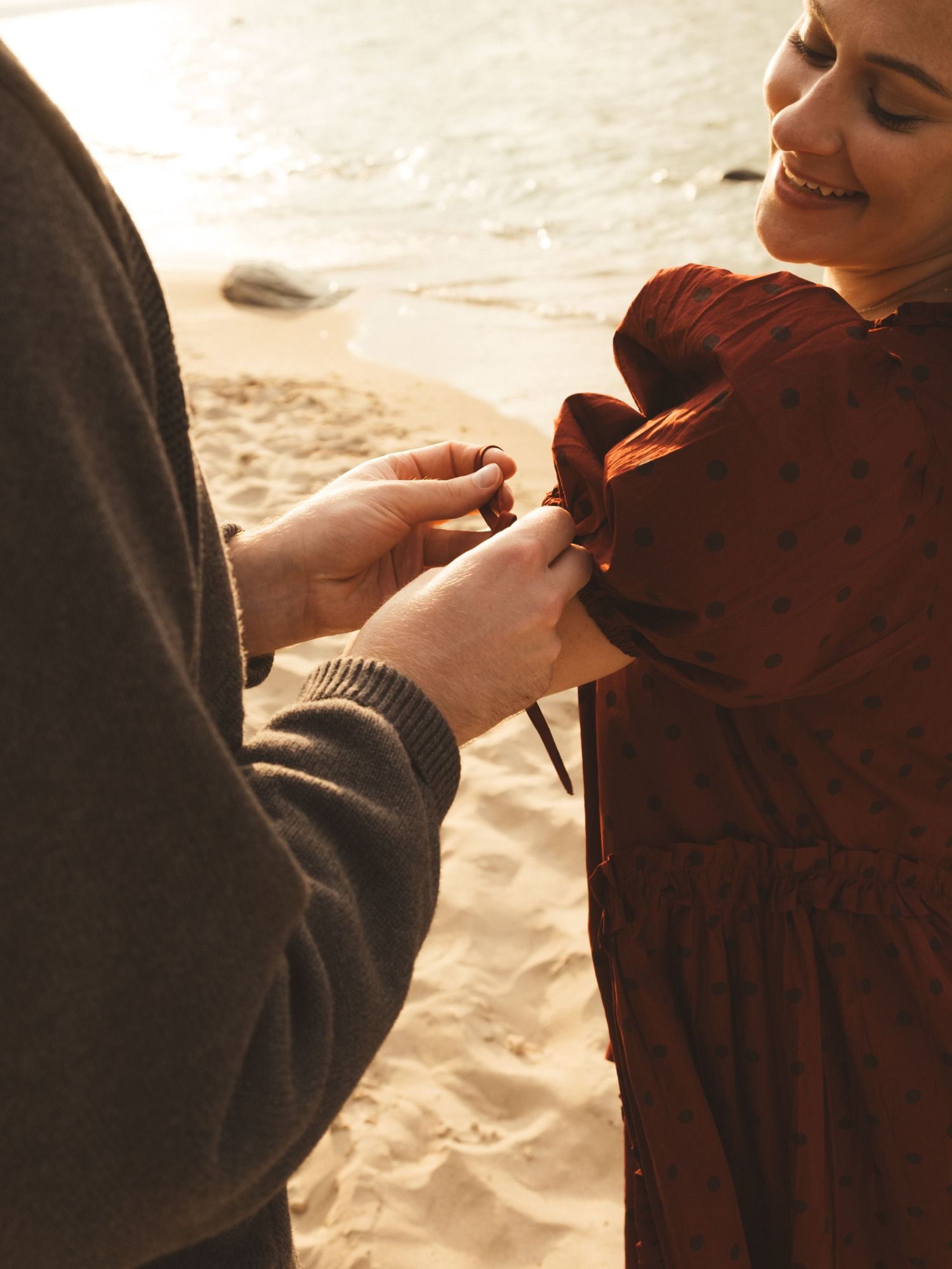 A man ties the bow of a woman's rust polka dot dress on a sandy beach at golden hour.