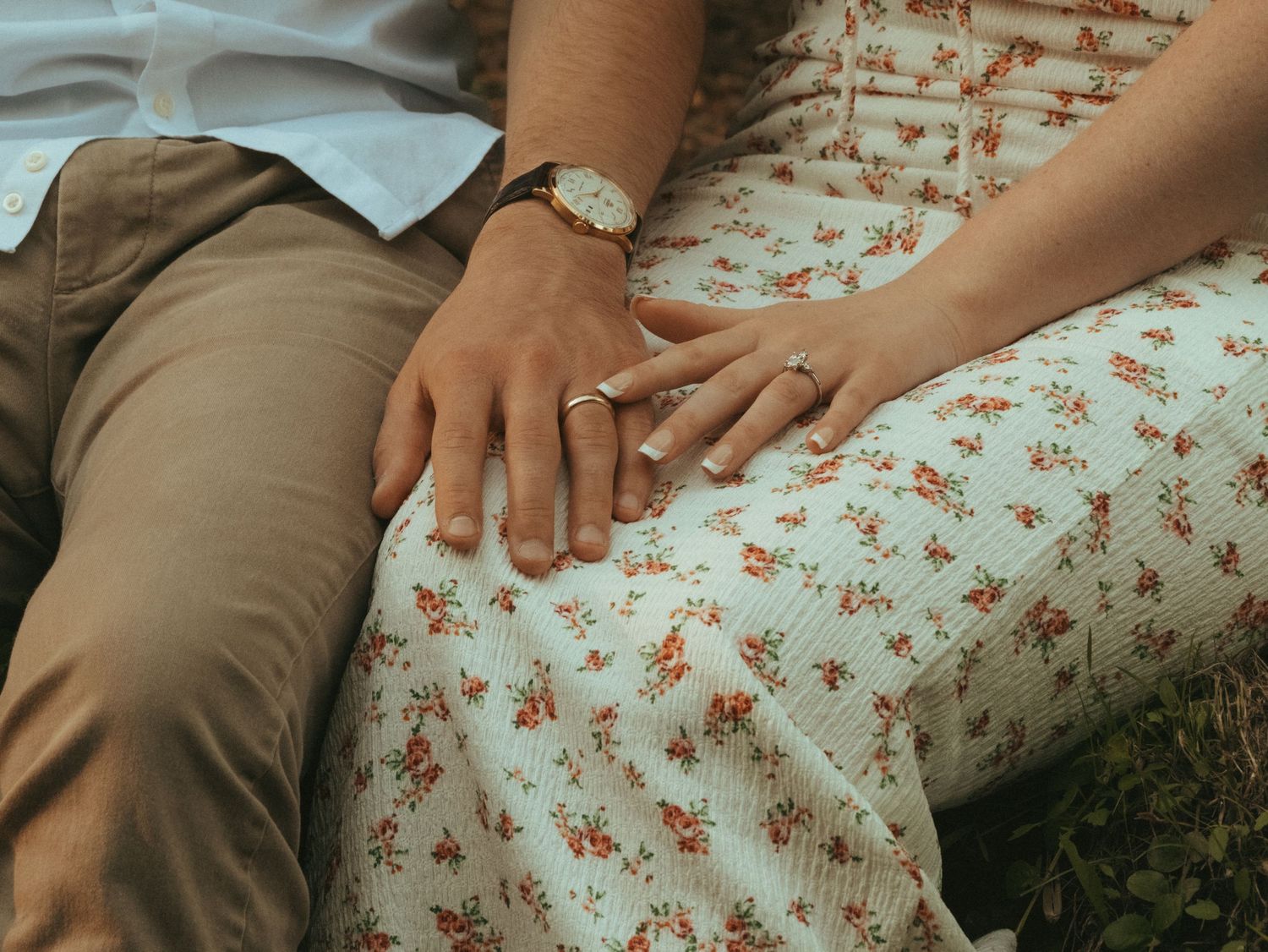 Close-up of a couple holding hands, wearing wedding rings, sitting together on grass in casual floral and tan outfits.