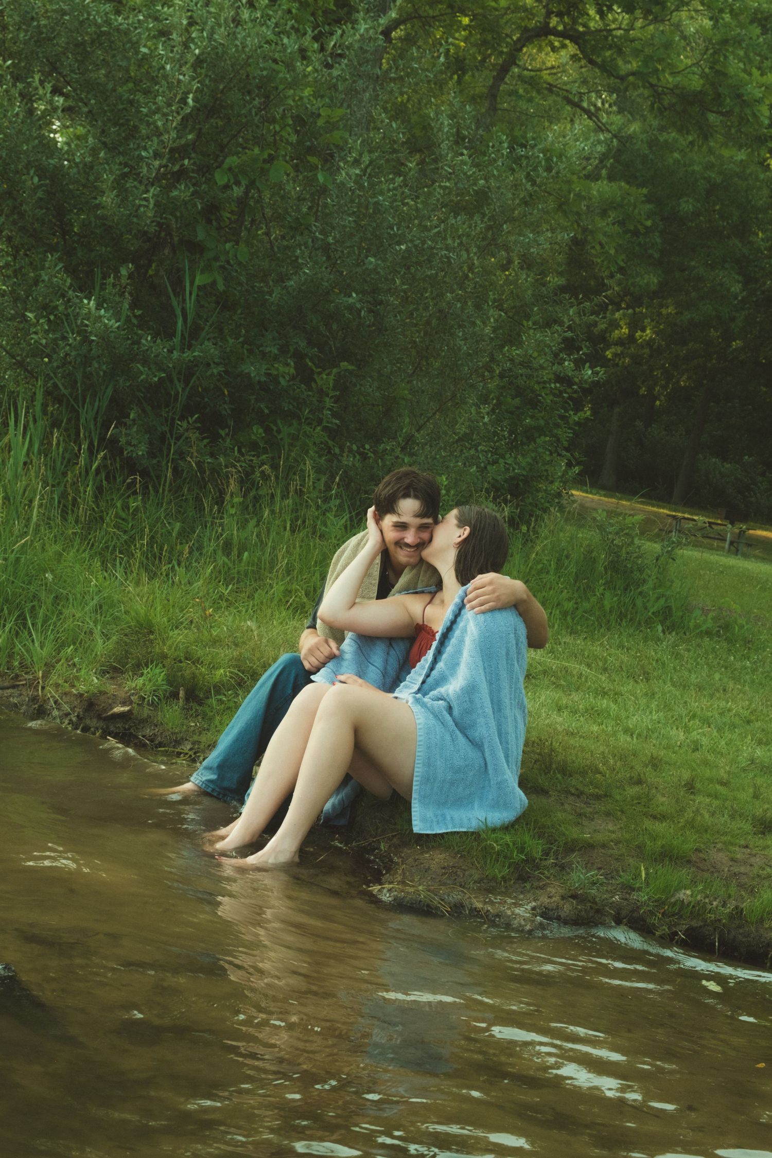 Couple sharing a romantic kiss by a serene river, wrapped in a blue blanket surrounded by lush green trees.