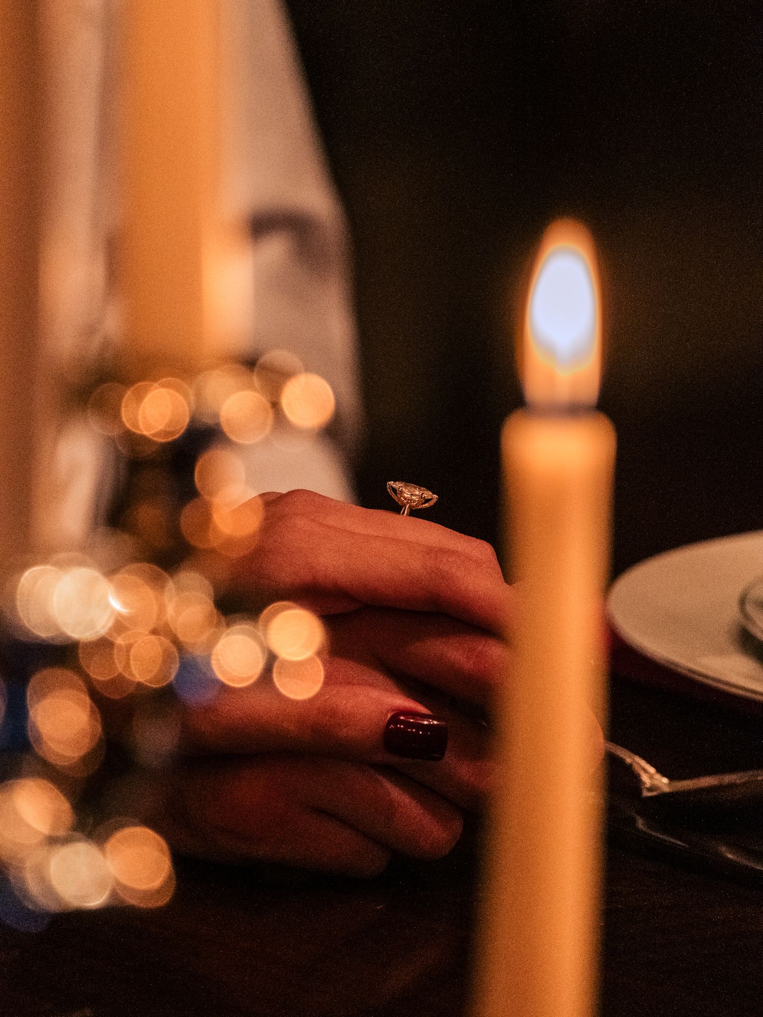A lit candle glows warmly beside hands with dark nail polish against a background of soft bokeh lights.