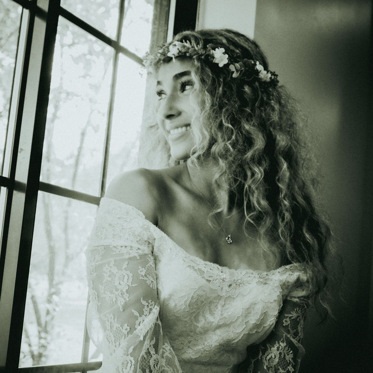 A dreamy black and white portrait of a bride with curly hair and floral crown looking out a window in vintage style.