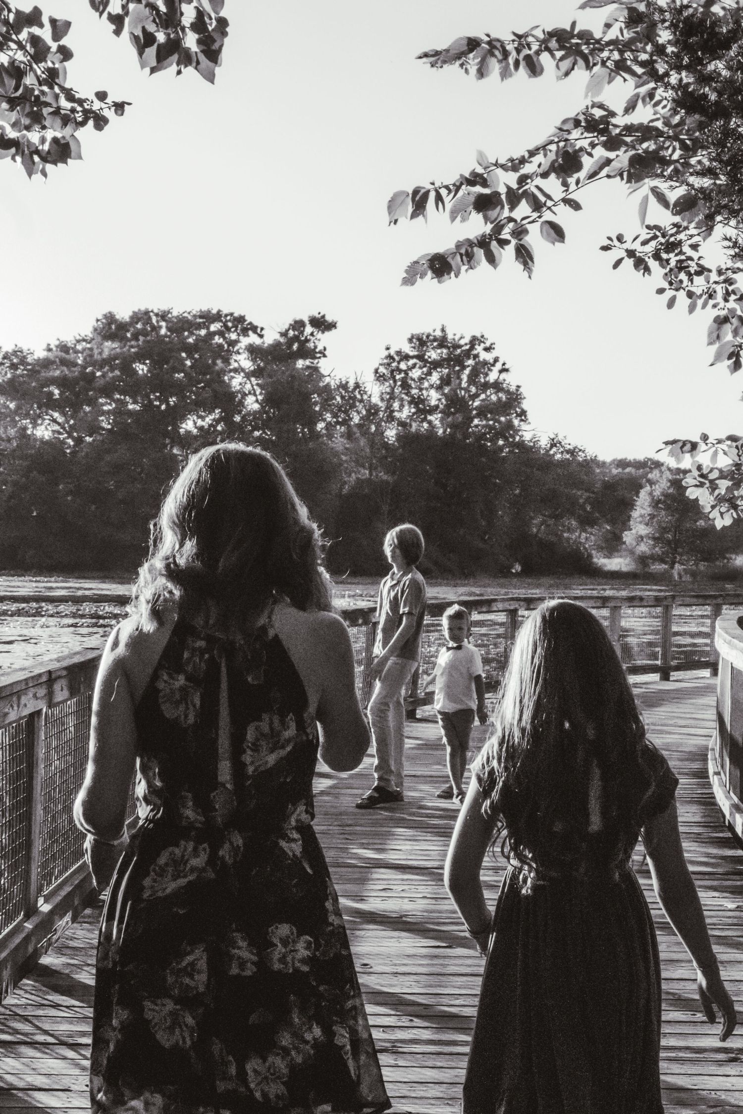 Line of people in dark clothing walking on a wooden boardwalk surrounded by trees in black and white.