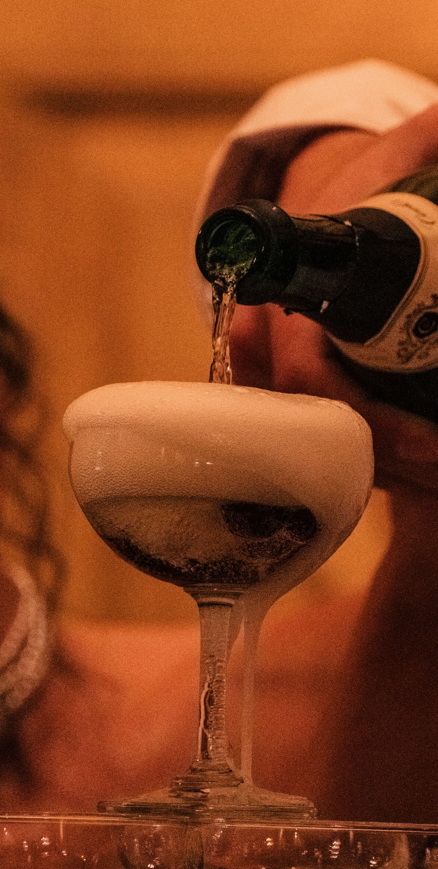 Champagne being poured into multiple coupe glasses in a dimly lit setting with warm amber lighting.