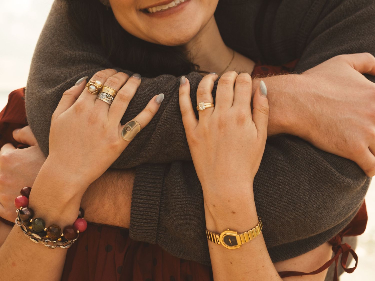 Couple embracing warmly, woman wearing gold watch, rings, and beaded bracelet, hands clasped around partner.