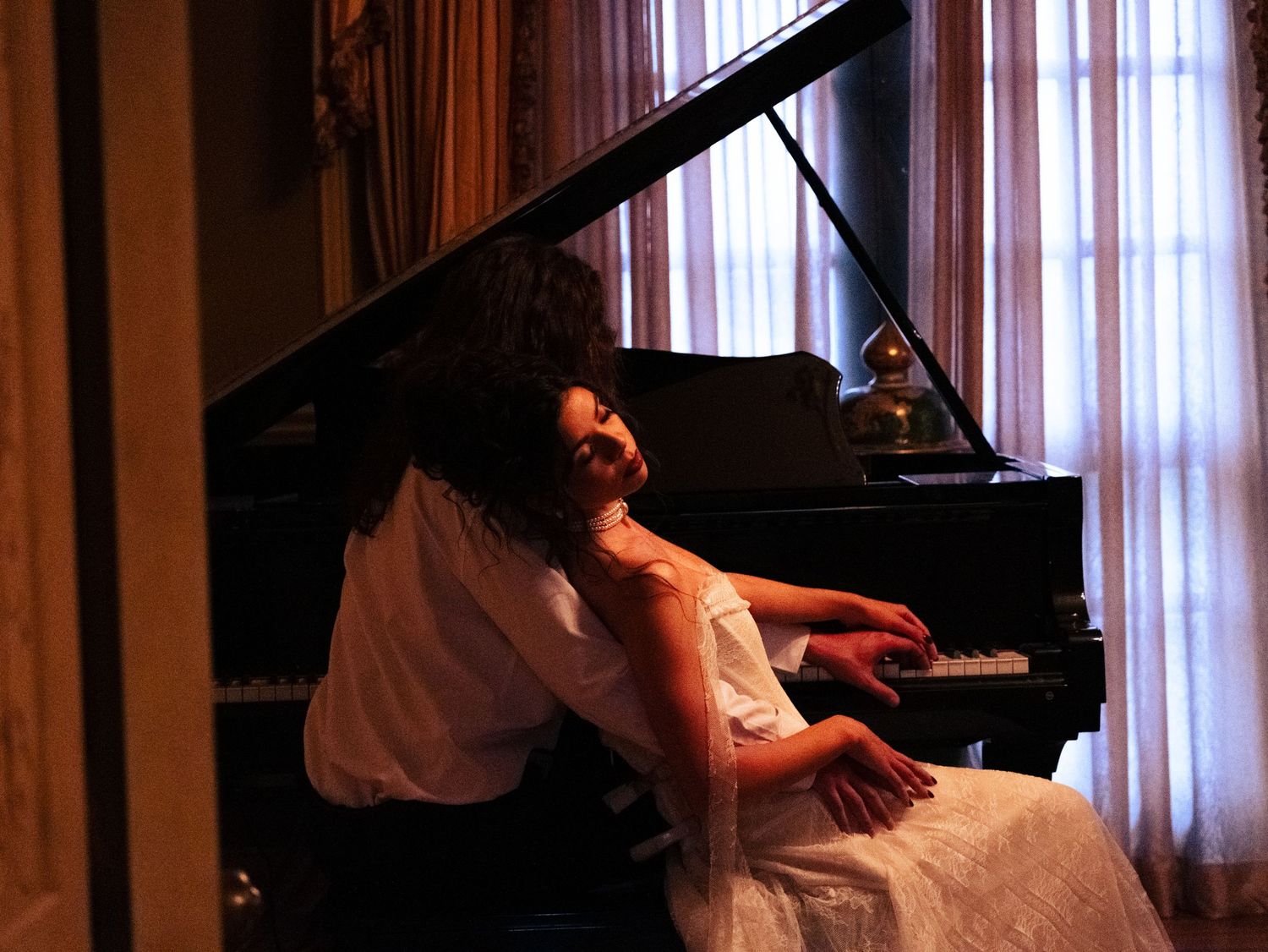 A person plays a grand piano in an elegant room with white curtained windows and warm lighting.