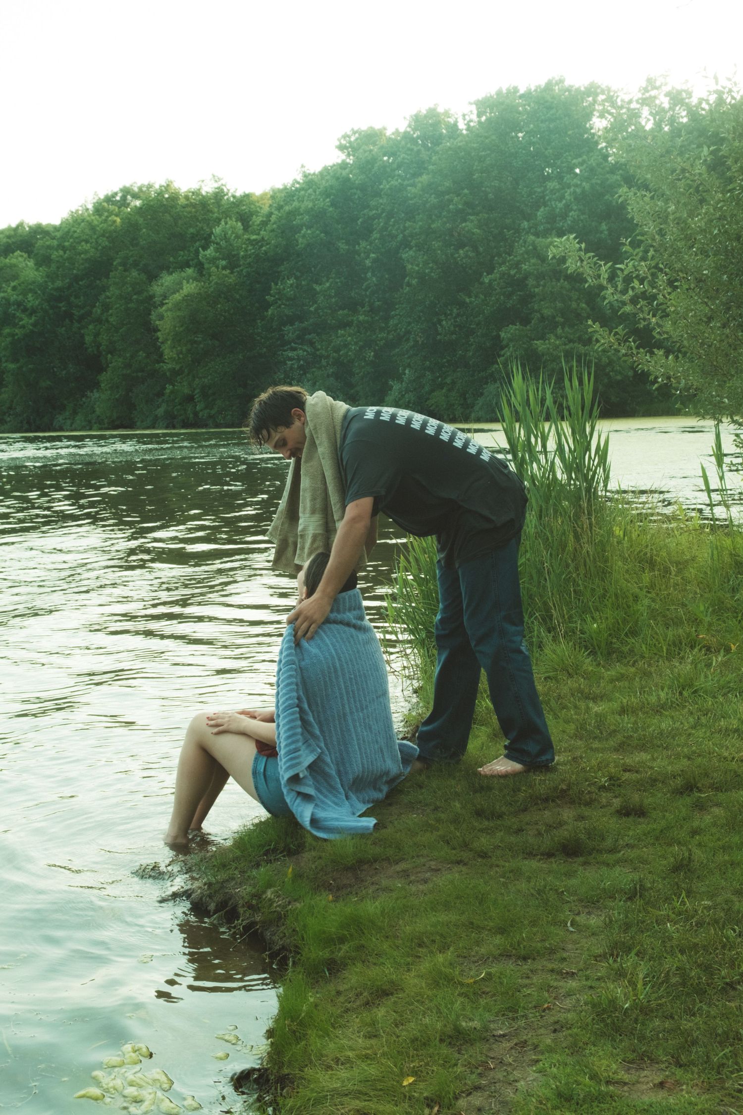 Man wrapping a blue towel around a woman sitting by a peaceful lakeside surrounded by lush green trees.