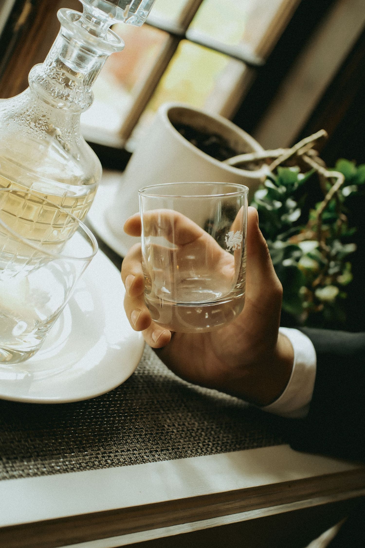 Hand in formal attire holding an empty crystal glass next to an ornate decanter on a textured surface.