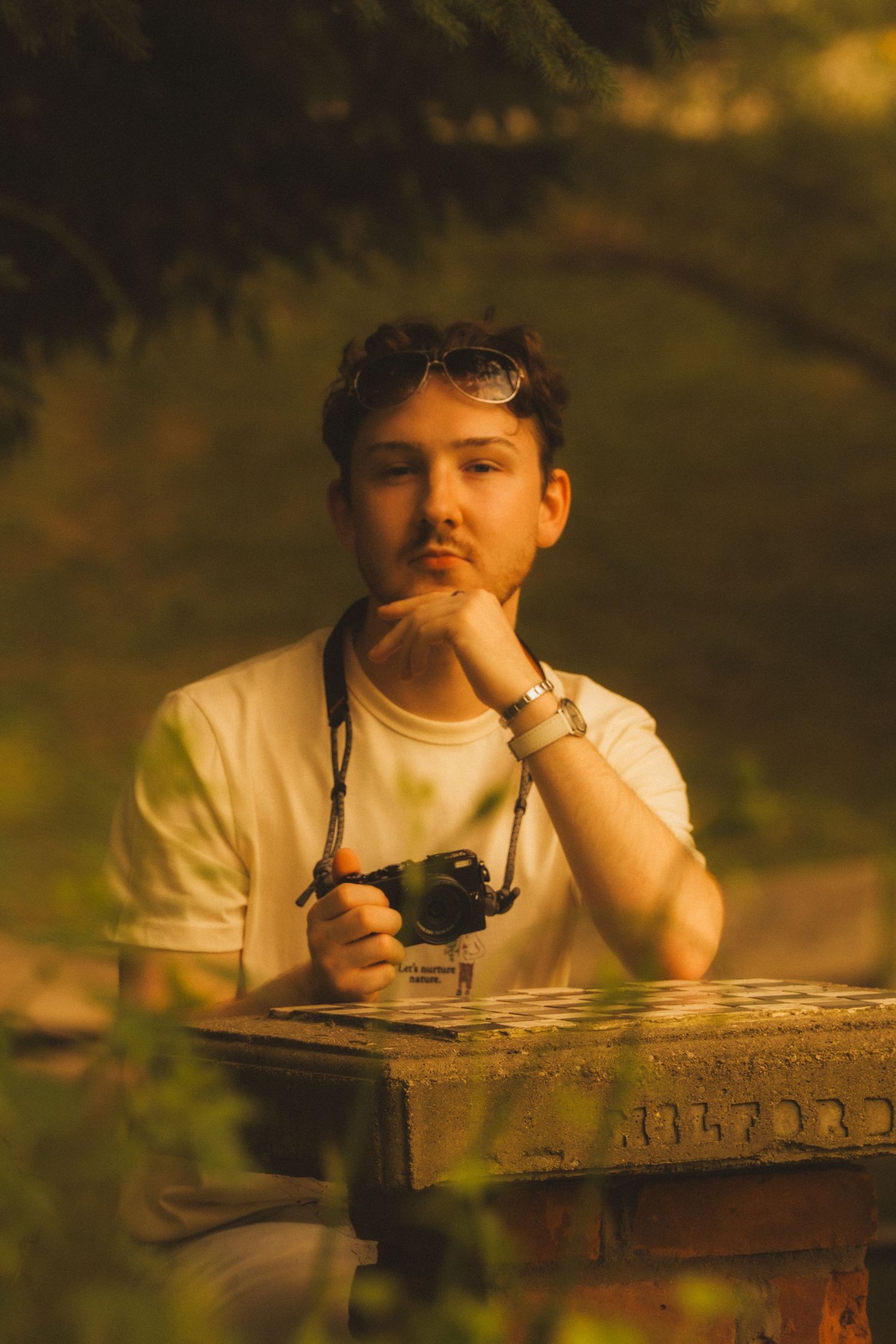 A person rests their head on a wooden railing at sunset while holding a camera in a contemplative outdoor scene.