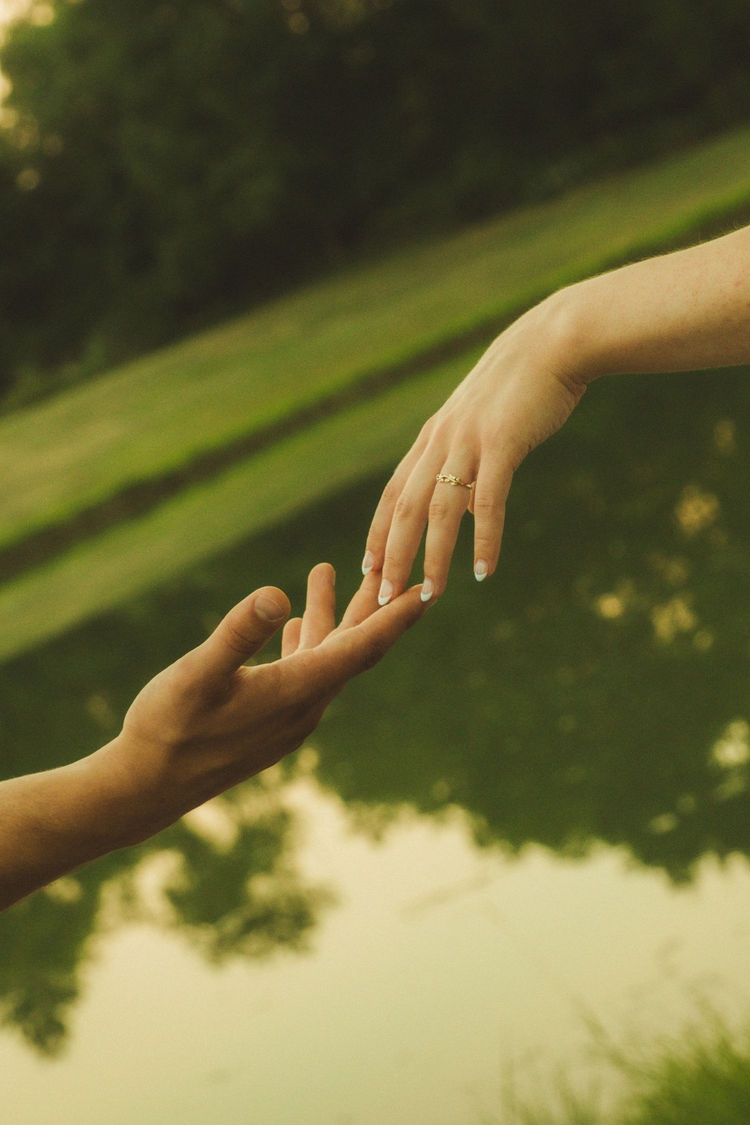 Two hands gently reaching toward each other outdoors, one wearing a delicate gold ring, with a green park background.