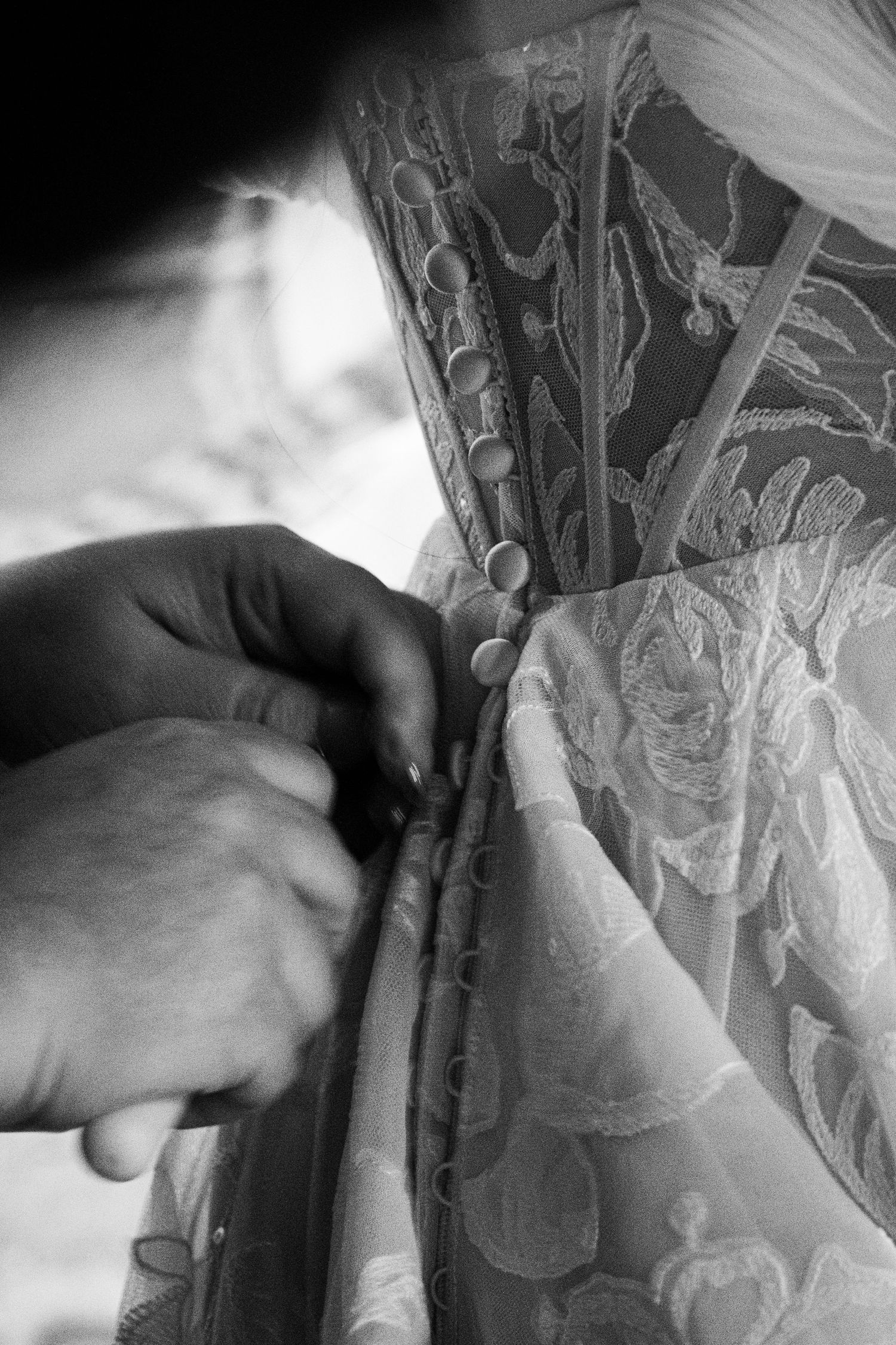 Black and white photo of hands fastening intricate lace buttons on an elegant bridal wedding gown.