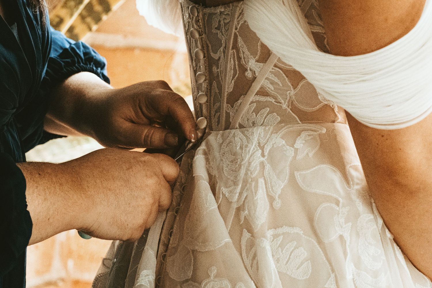 Close-up shot of hands buttoning up a delicate white lace wedding dress with intricate floral patterns.