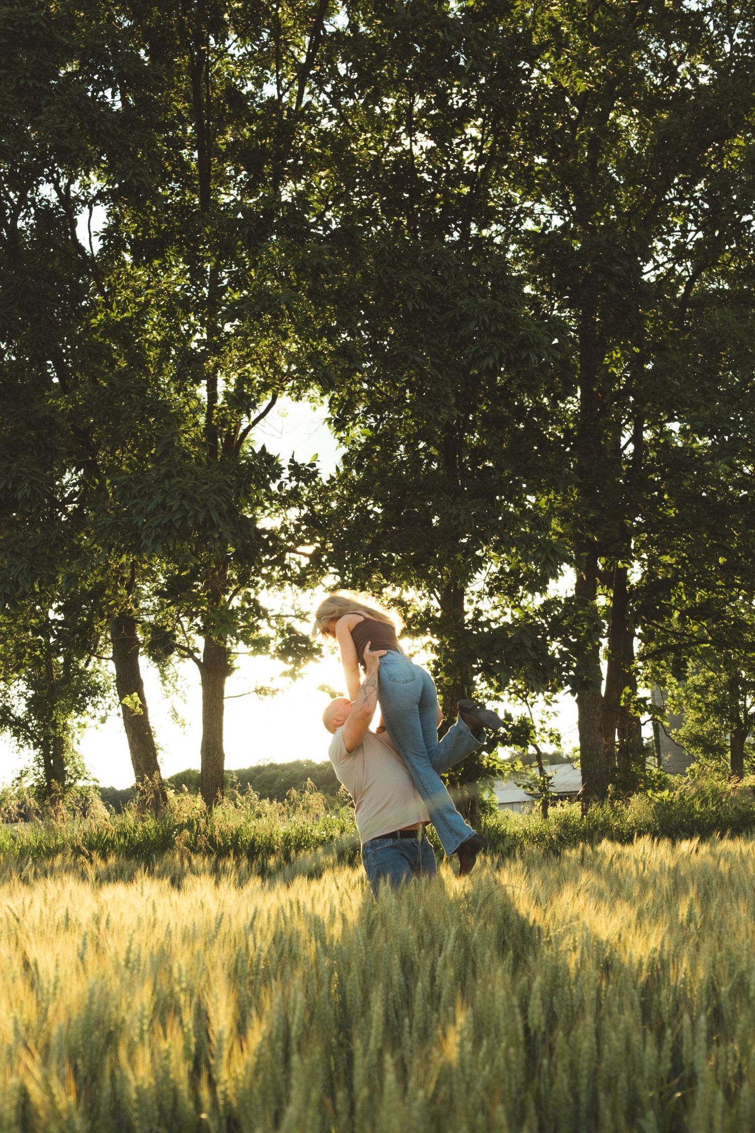 A person lifts a child joyfully in a sunlit wheat field surrounded by tall green trees.
