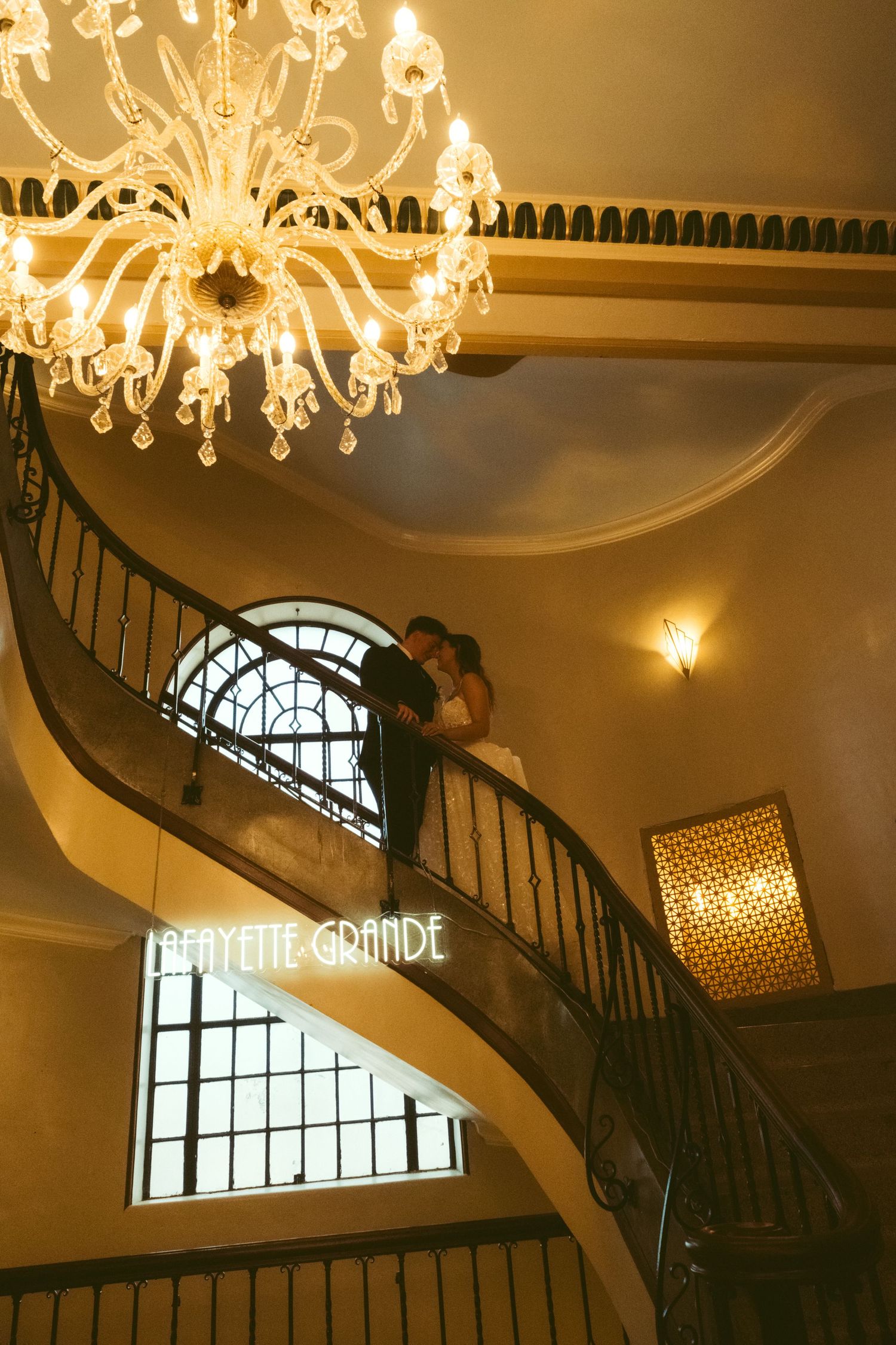 Couple embracing on elegant curved staircase beneath a grand chandelier at Lafayette Grande venue.