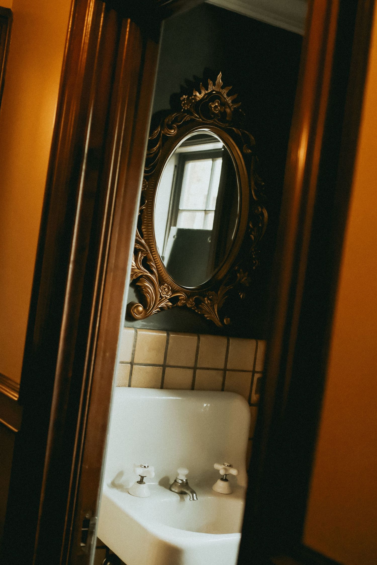 Ornate gold oval mirror above a white pedestal sink in a vintage bathroom with dark walls and warm wooden trim.