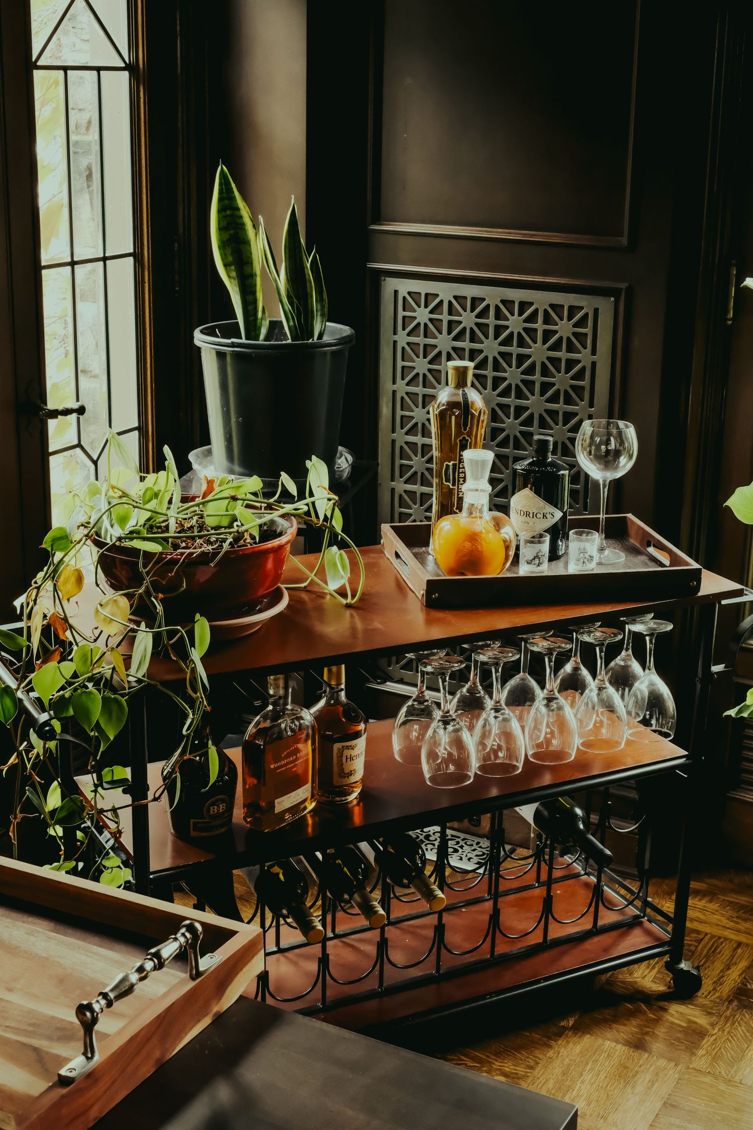 Elegant bar cart with wine glasses, liquor bottles, and houseplants in a dark, moody interior setting.