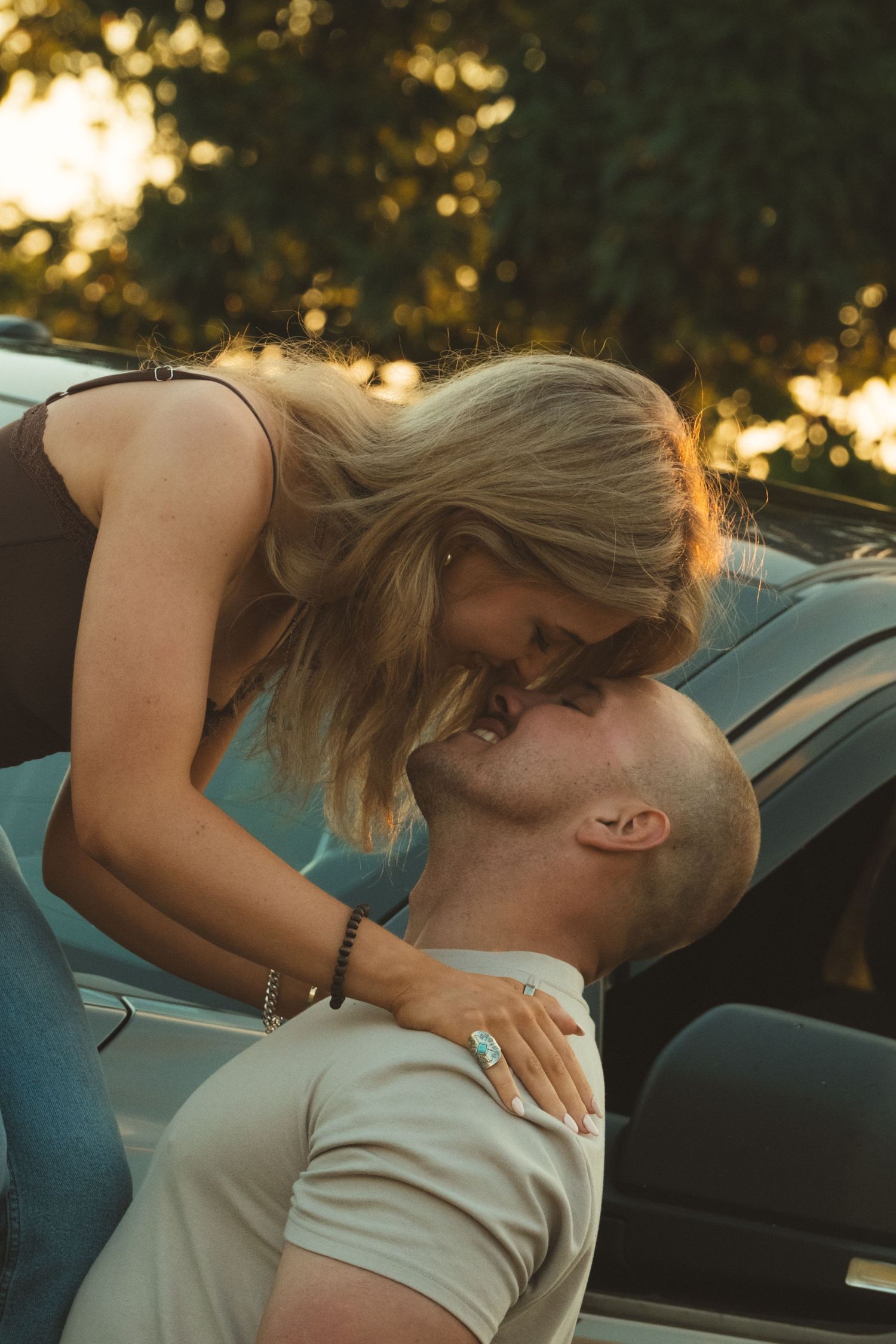 A couple shares a romantic kiss near a car outdoors, with golden sunlight filtering through lush green trees in the background.