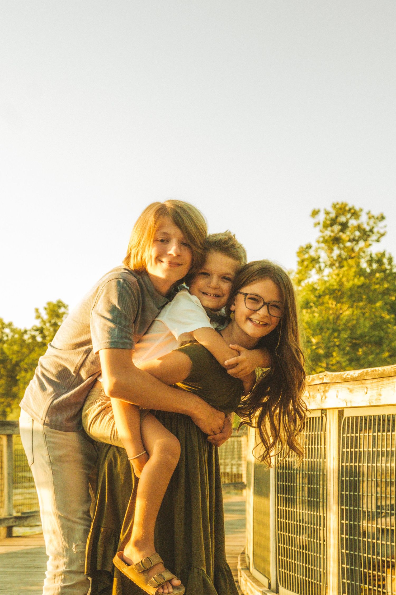 Three friends share joyful embraces while posing together at sunset along a fence during golden hour.