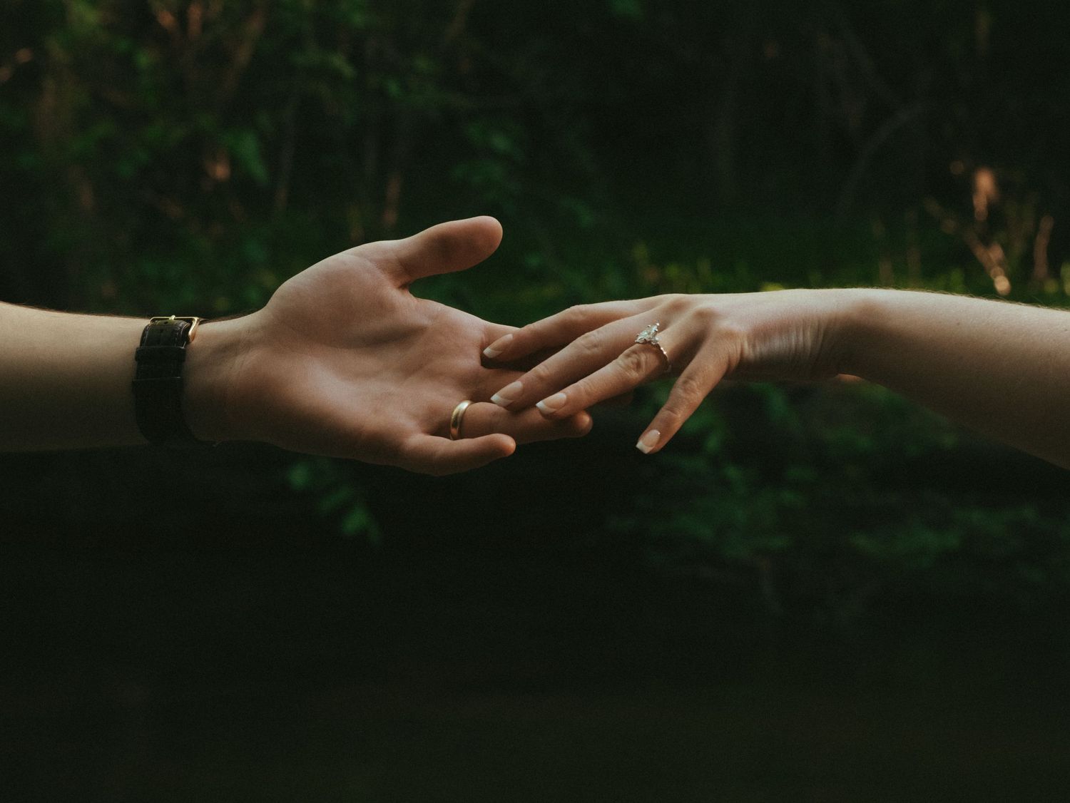 Two hands gently reaching toward each other, one wearing a ring, against a dark green background.