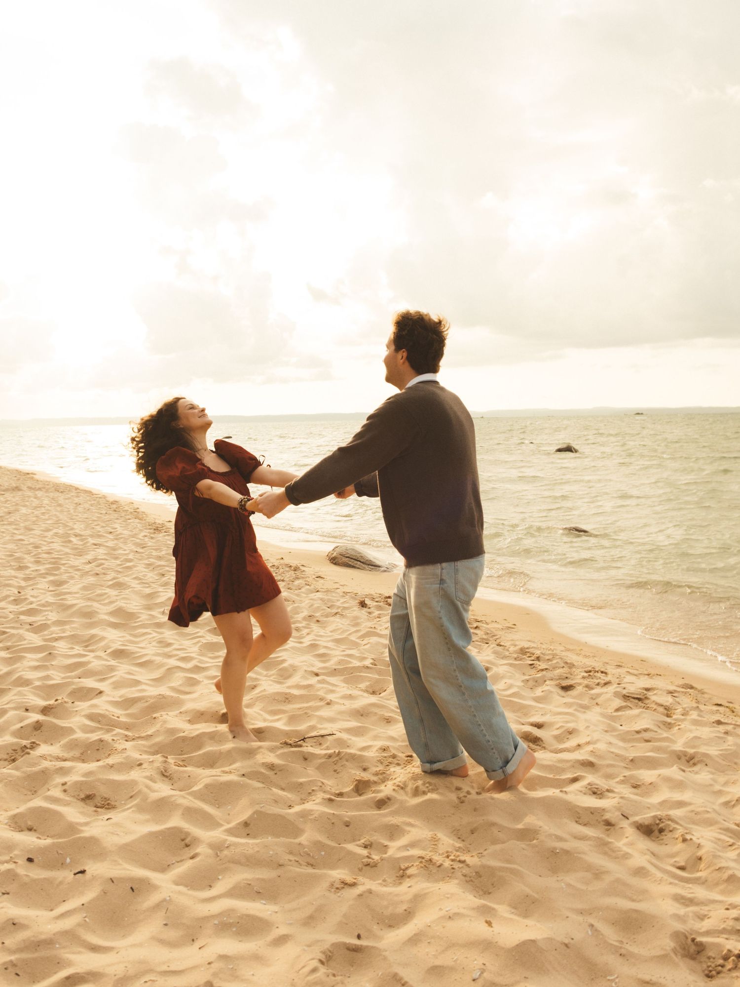 Couple dancing barefoot on sandy beach, holding hands and spinning joyfully near ocean waves under cloudy sky.