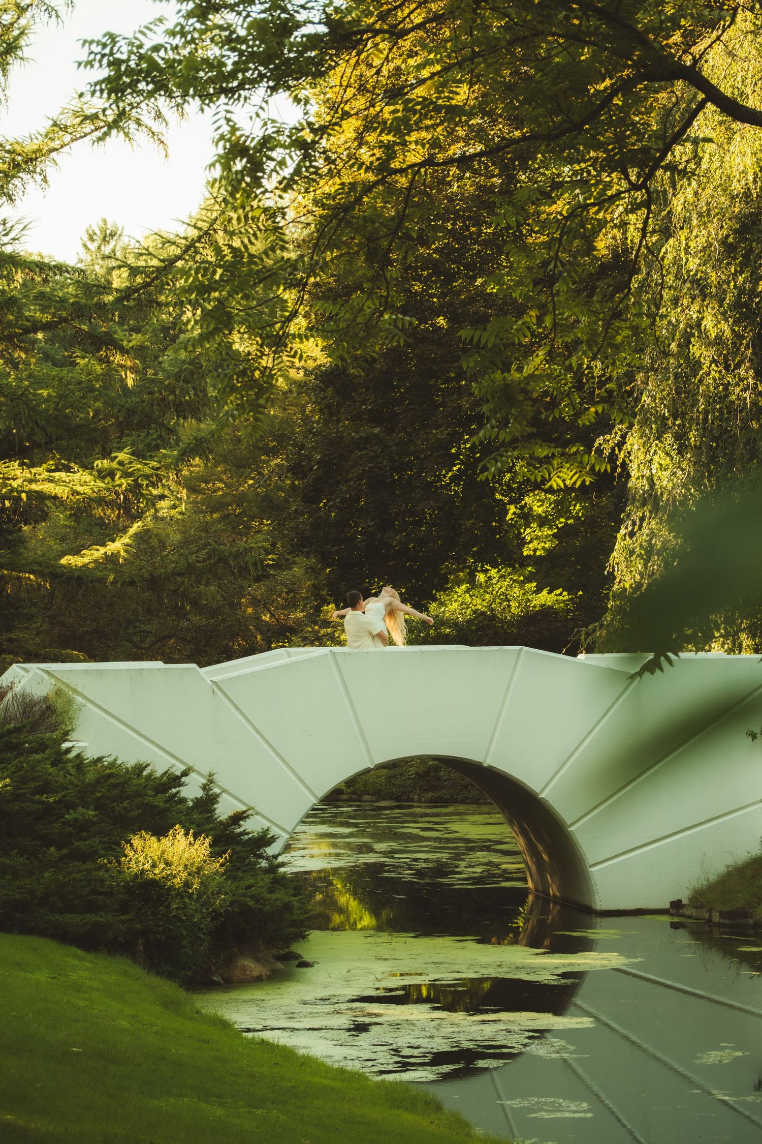 Couple in wedding attire stands on a white arched bridge over a lily pond surrounded by lush green trees in a park.