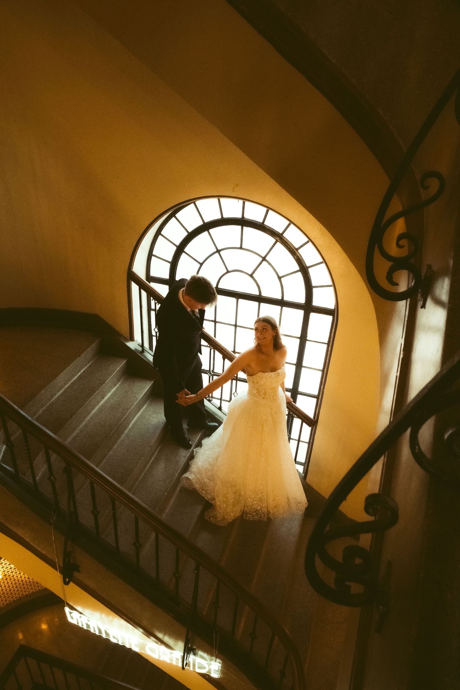 Bride and groom holding hands on elegant spiral staircase with arched window, captured from above in warm golden light.