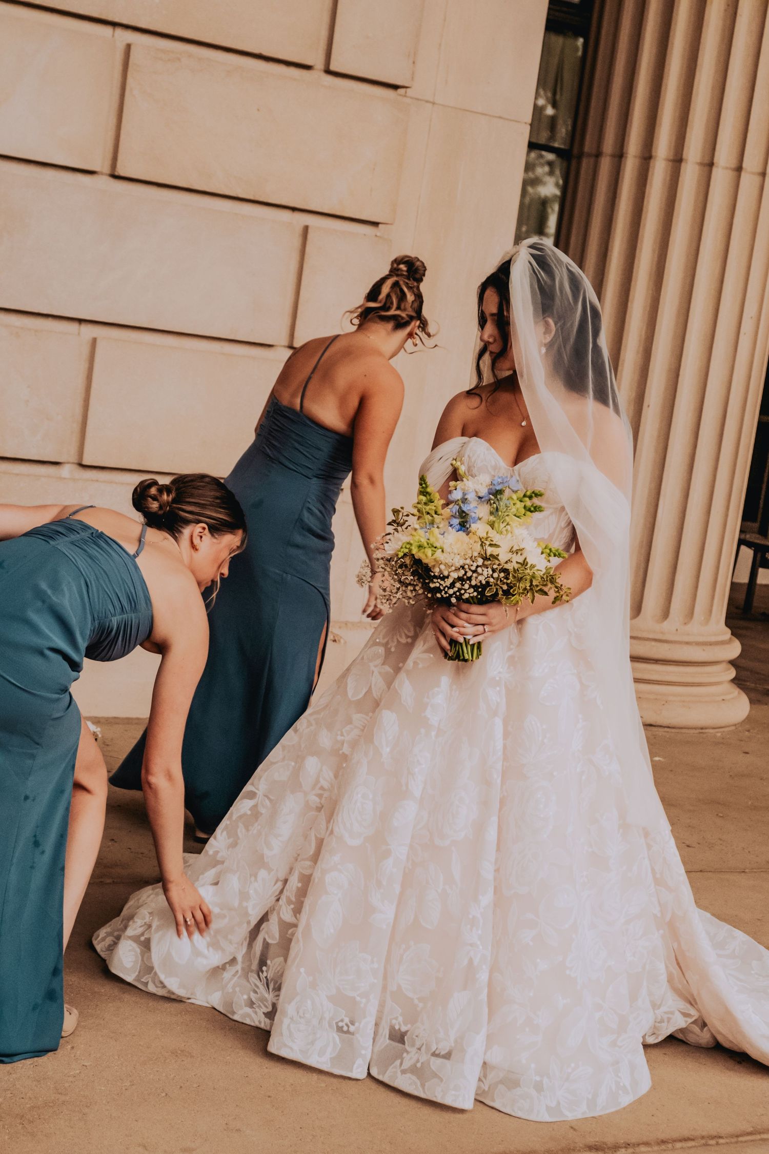 A bride in a white wedding gown holds a bouquet while attendants in teal dresses help with her dress near stone columns.