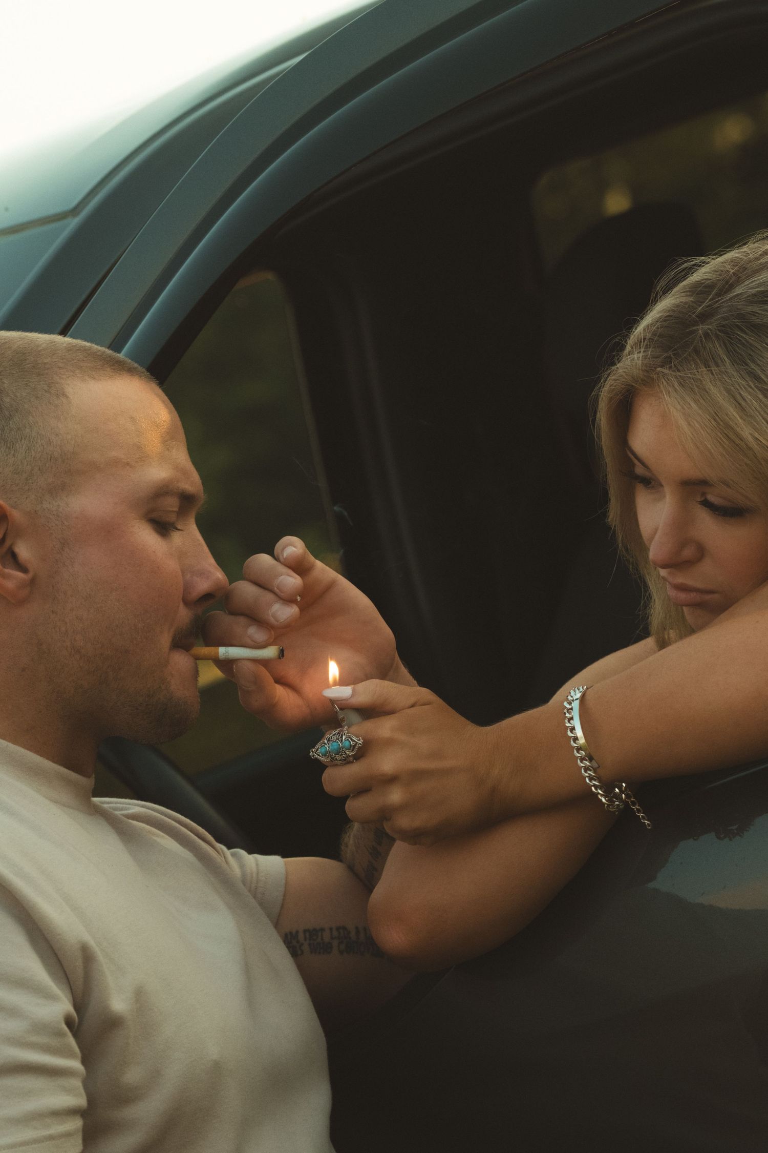 A woman lights a cigarette for a man sitting in a car window, wearing a bracelet and ring.