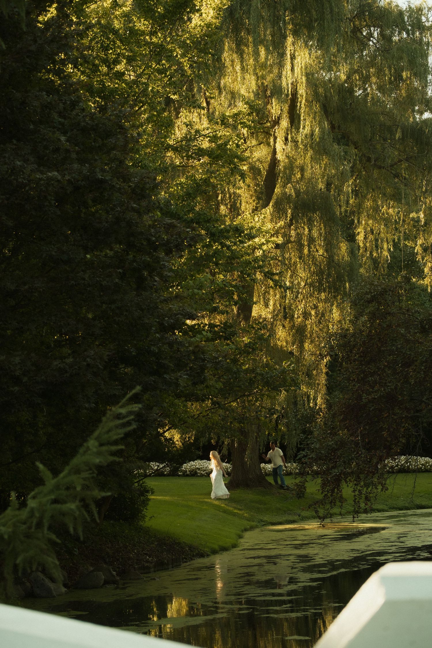 Bride in white dress stands near a reflective pond surrounded by lush green trees in golden sunlight.