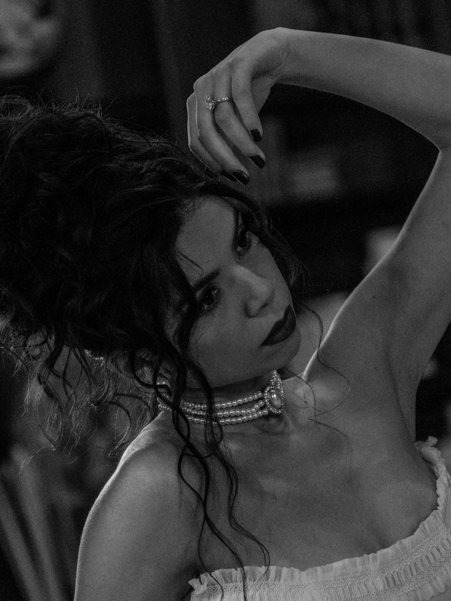A dramatic black and white portrait shows a figure in pearls with dark curly hair posing with raised arm against dark backdrop.
