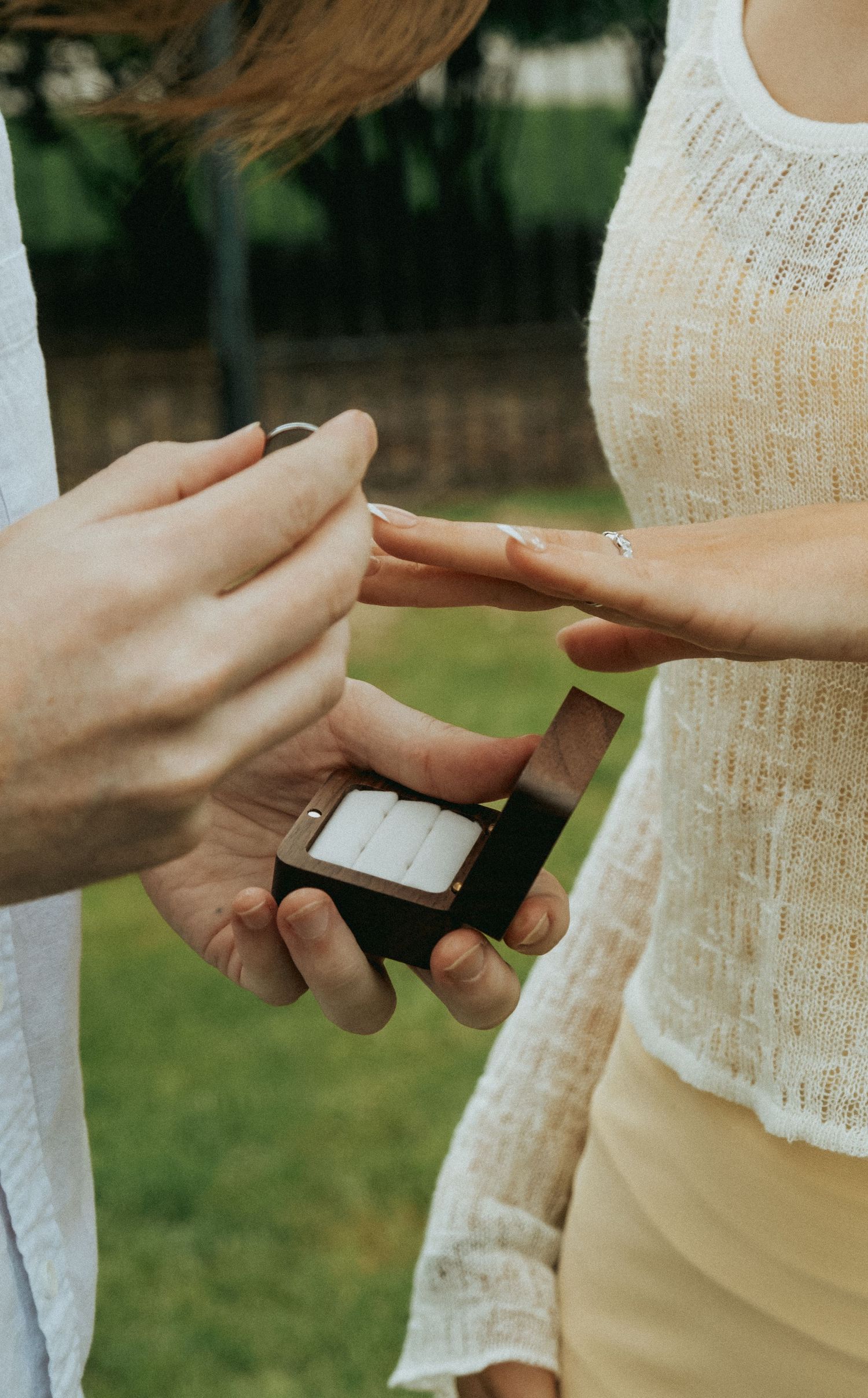 Close-up of hands exchanging a ring during an outdoor engagement proposal with white lace clothing visible.