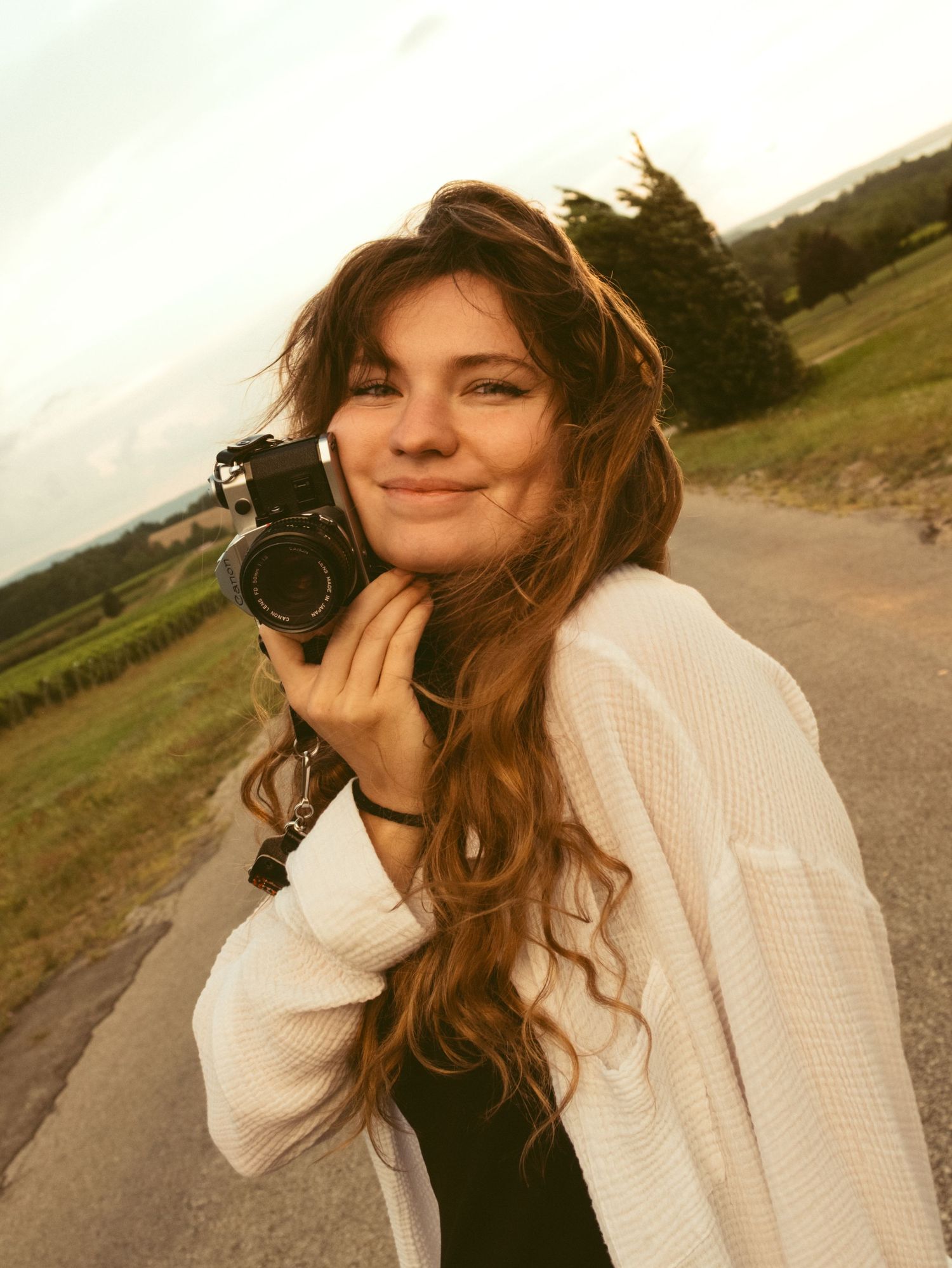 Young woman with long brown hair smiles while holding a vintage camera outdoors on a rural road at dusk.