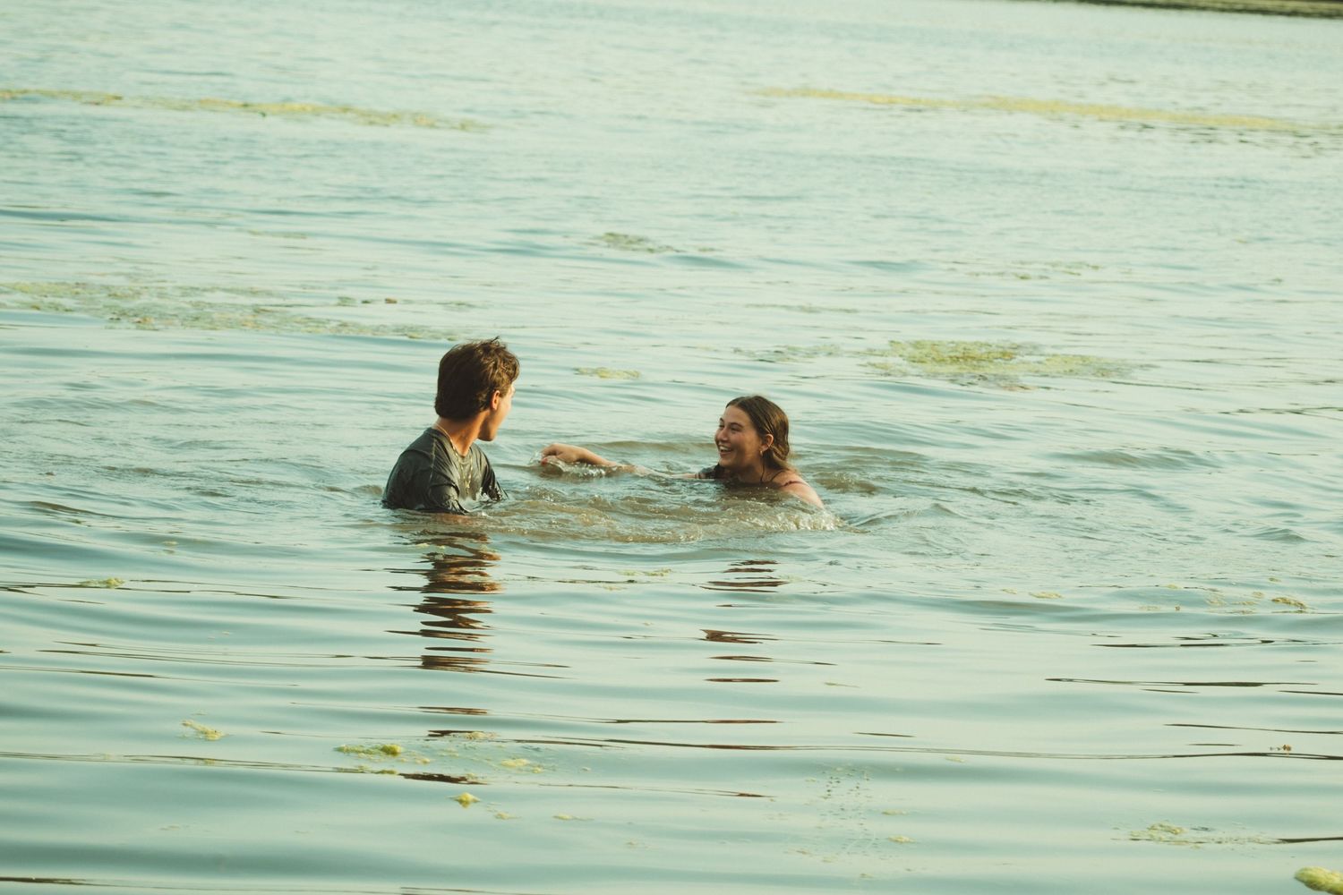 Two people swimming and talking in calm open water on a sunny day.