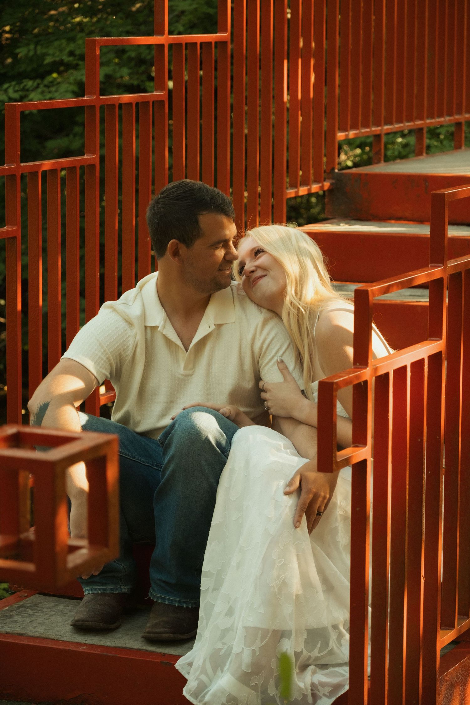 A couple sits together on red wooden steps, sharing a tender moment, with the woman in a white dress leaning toward the man.