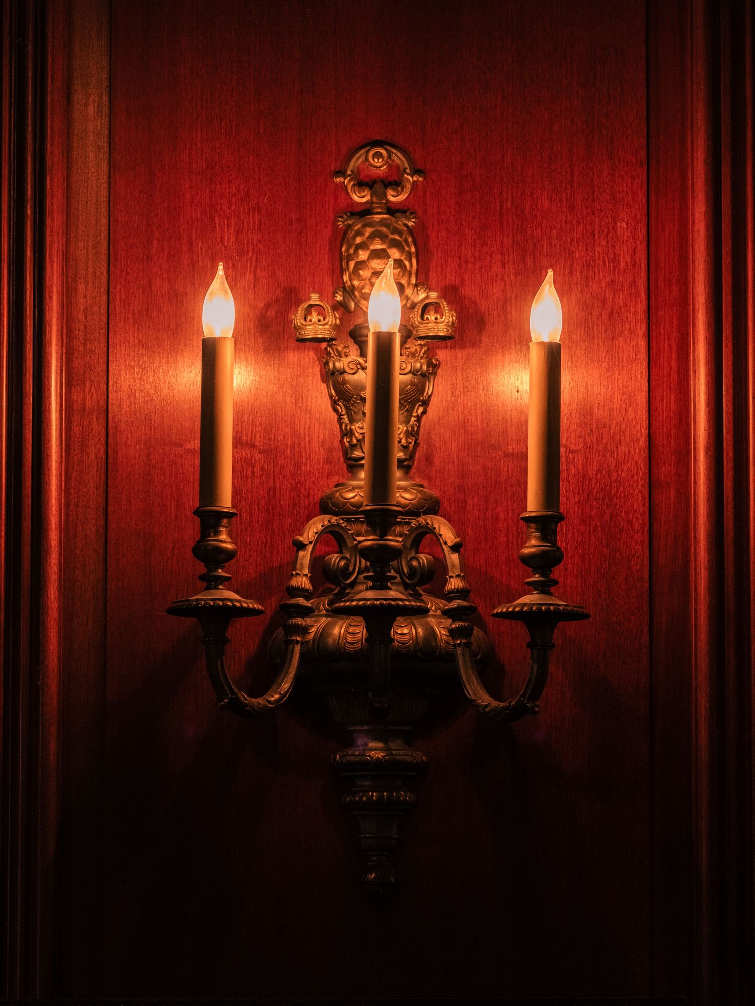 Ornate antique wall sconces with candle-style lights cast a warm glow against deep red wallpaper in dramatic lighting.