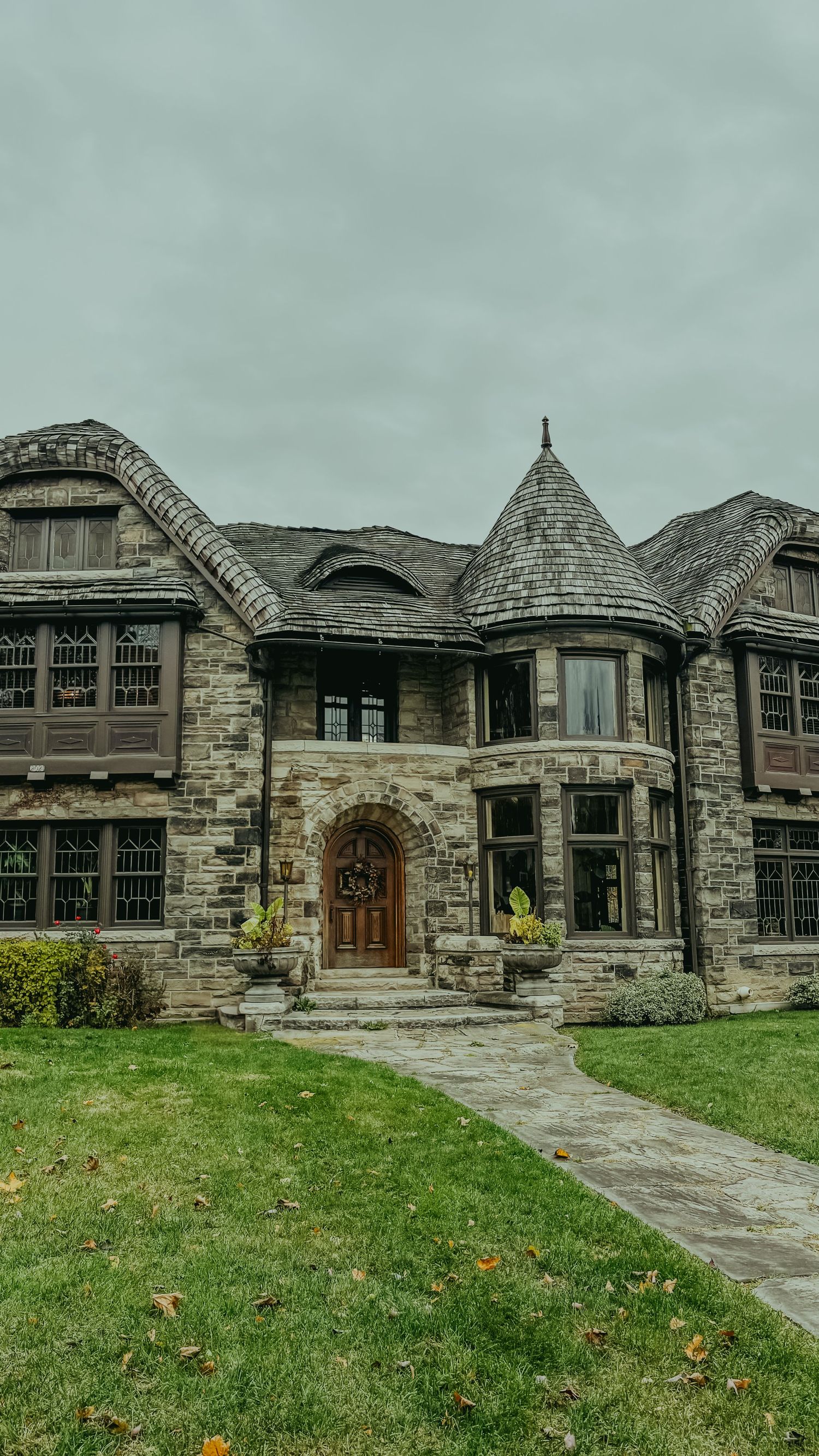 A grand stone Tudor-style mansion with turret, arched wooden door, leaded windows, and manicured lawn under overcast skies.
