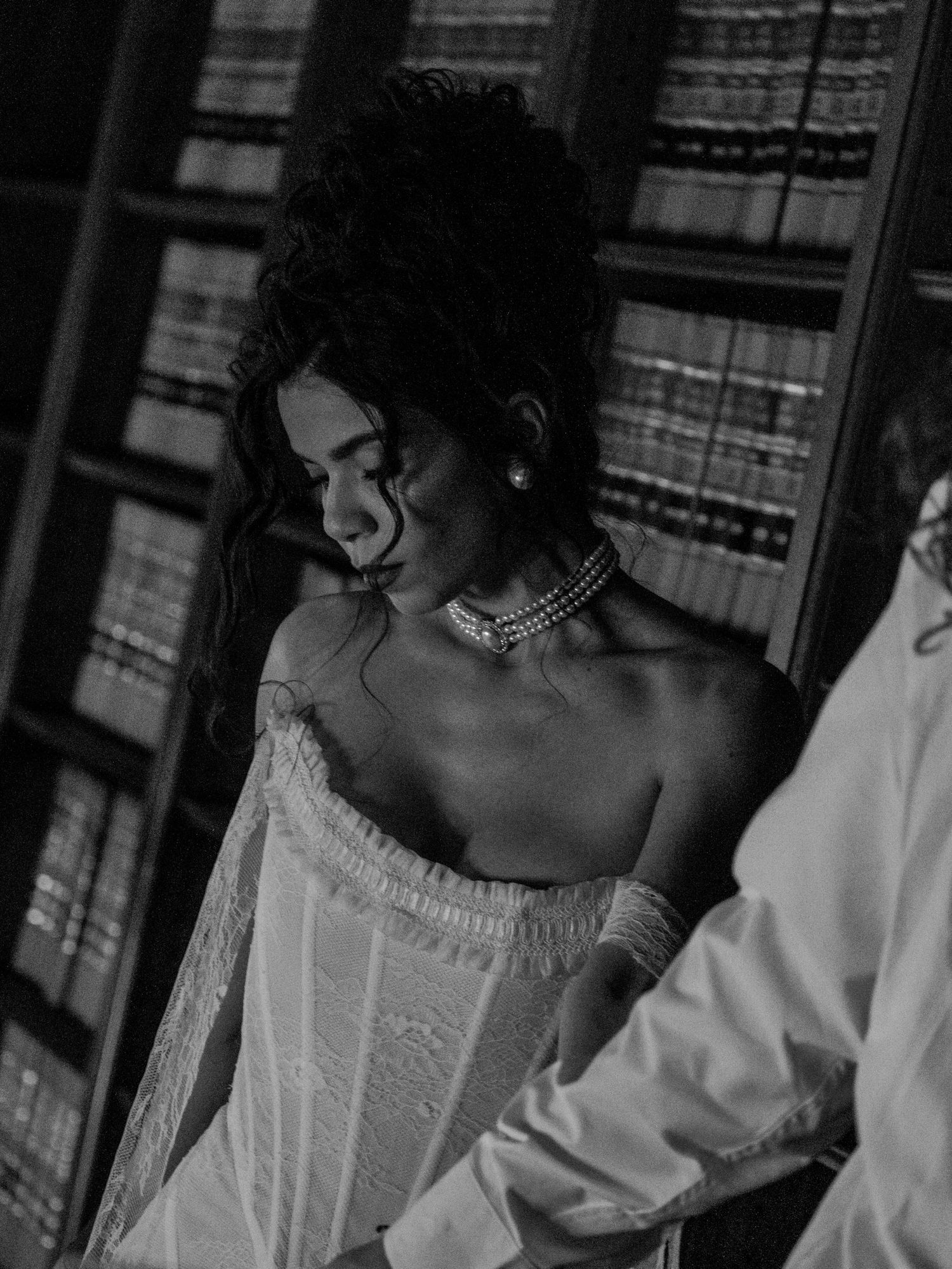 A black and white photograph of a person in a white dress gazing to the side with bookshelves in the background.