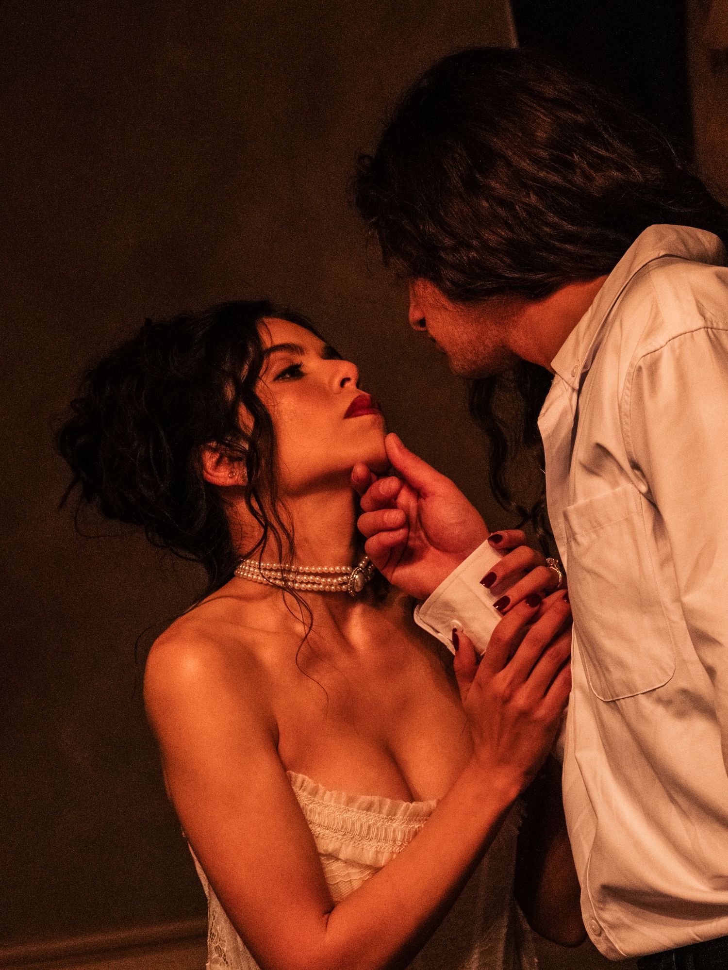 Romantic couple in intimate moment with woman in white dress and pearl necklace gazing up at man in dramatic lighting.
