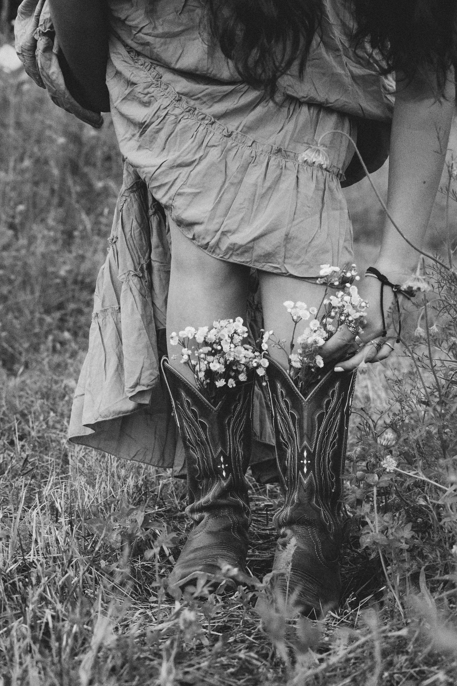 Black and white photo of woman in cowboy boots and floral dress holding wildflowers tucked into her boots outdoors.