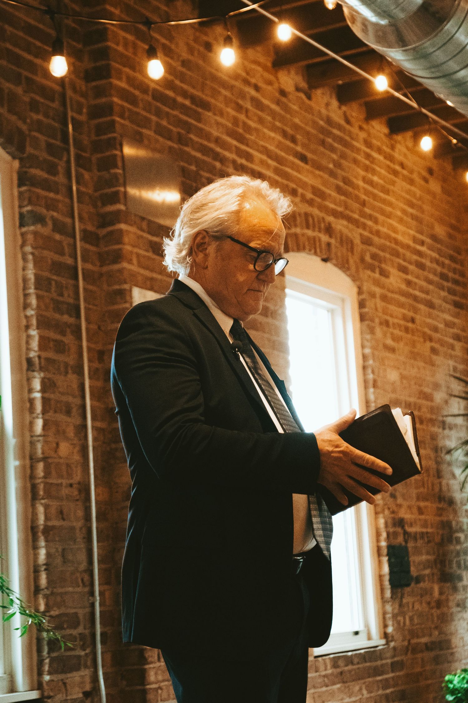 A person in a dark suit reads from a book inside a rustic brick-walled venue with string lights overhead.