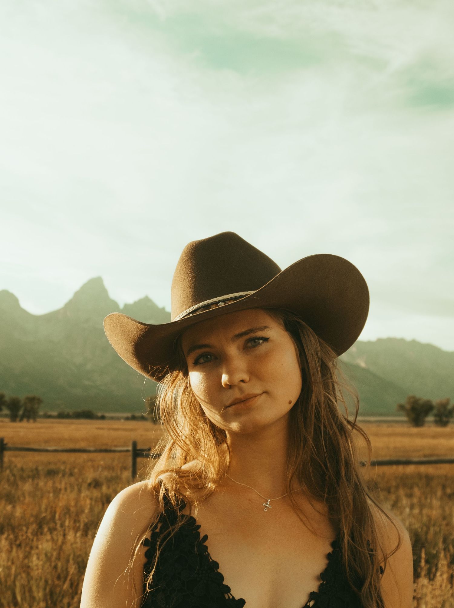 A person in a cowboy hat poses in a field during golden hour with mountains visible in the background.