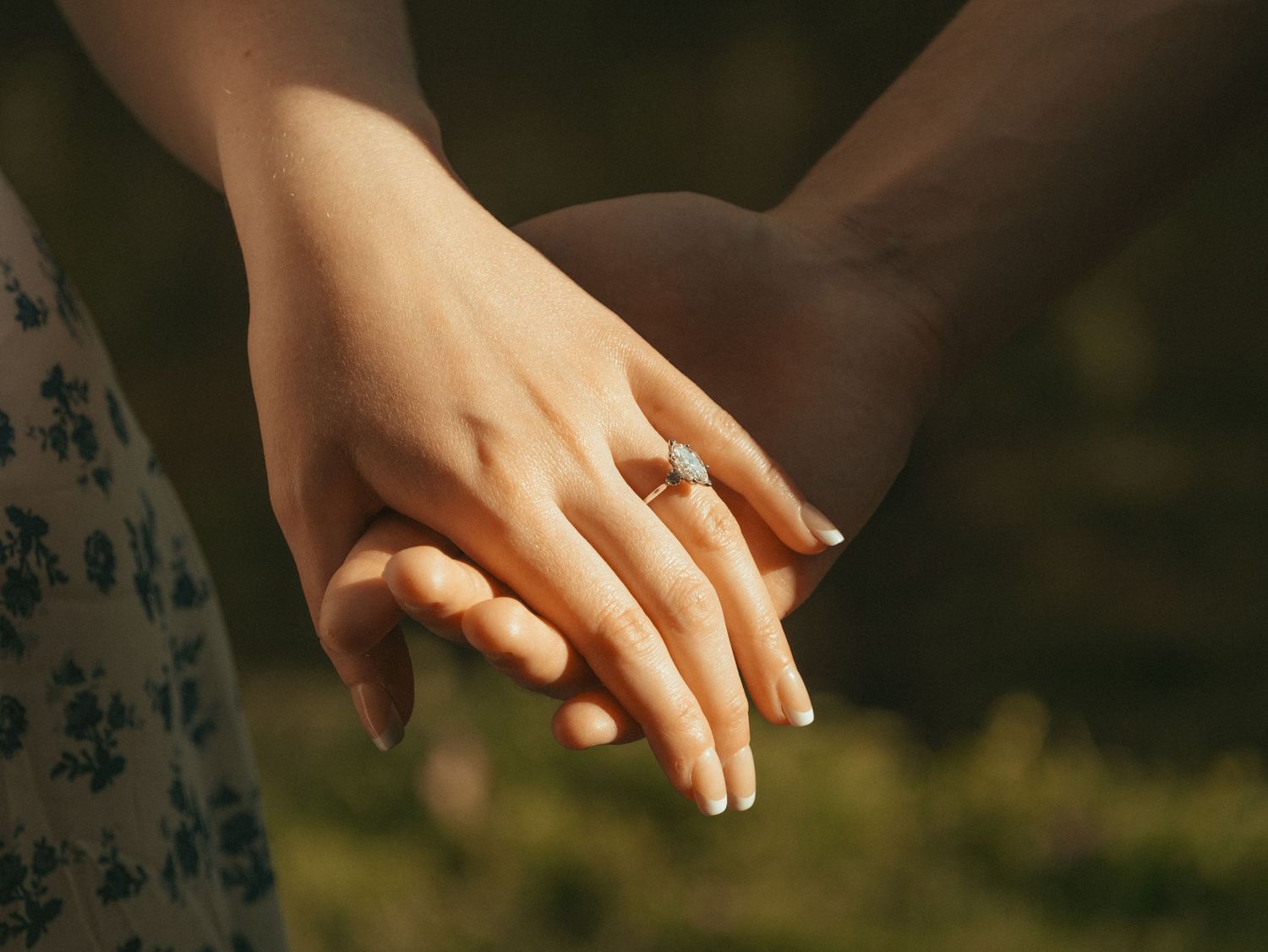 Close-up of two people holding hands, woman wearing a diamond engagement ring, romantic outdoor setting.